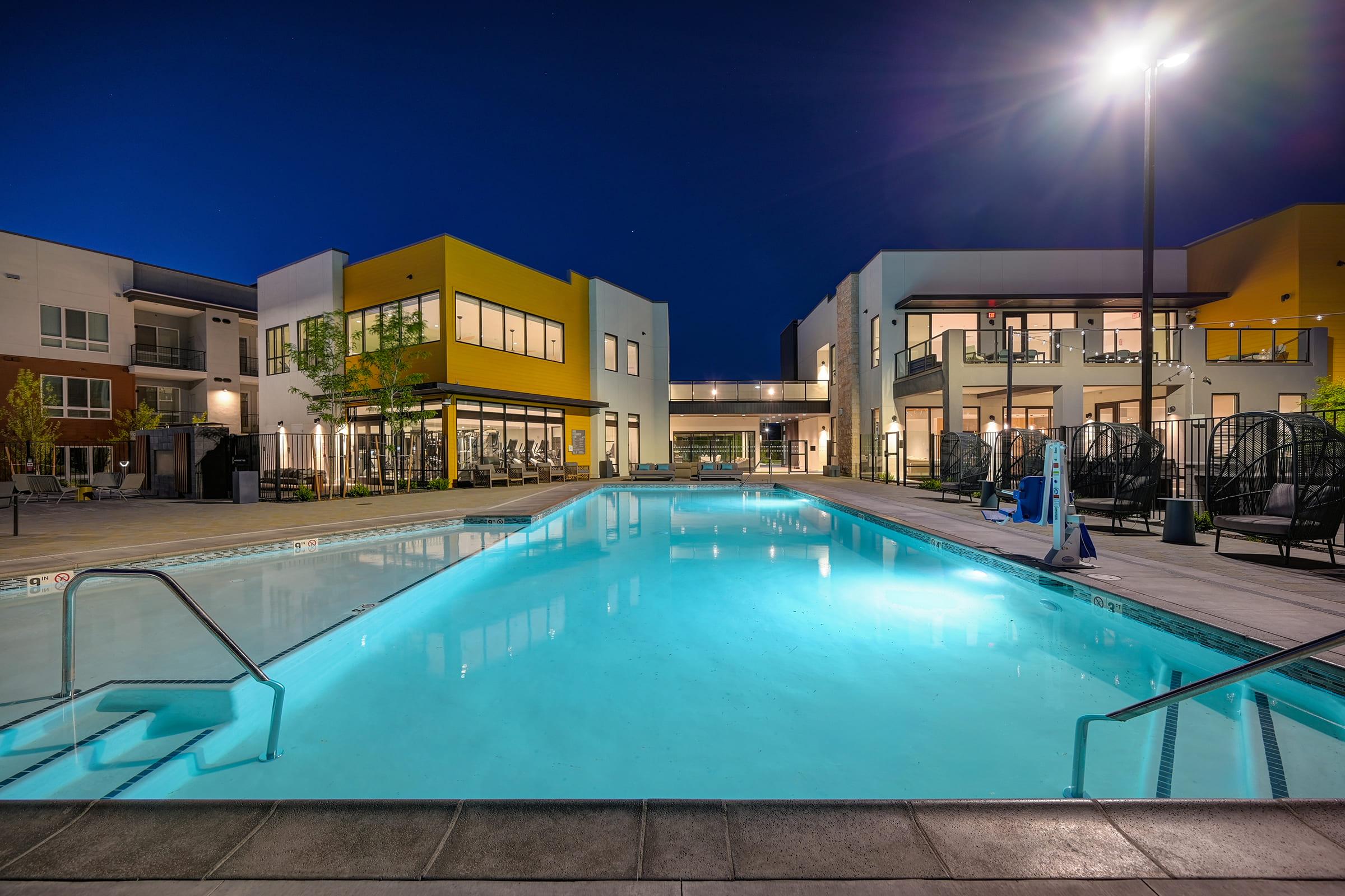 A serene pool area at night, illuminated by soft lights. The water glows blue, reflecting the surrounding buildings with modern architecture. Lounge chairs are positioned along the poolside, and greenery adds a touch of nature. The overall atmosphere is calm and inviting, perfect for relaxation.