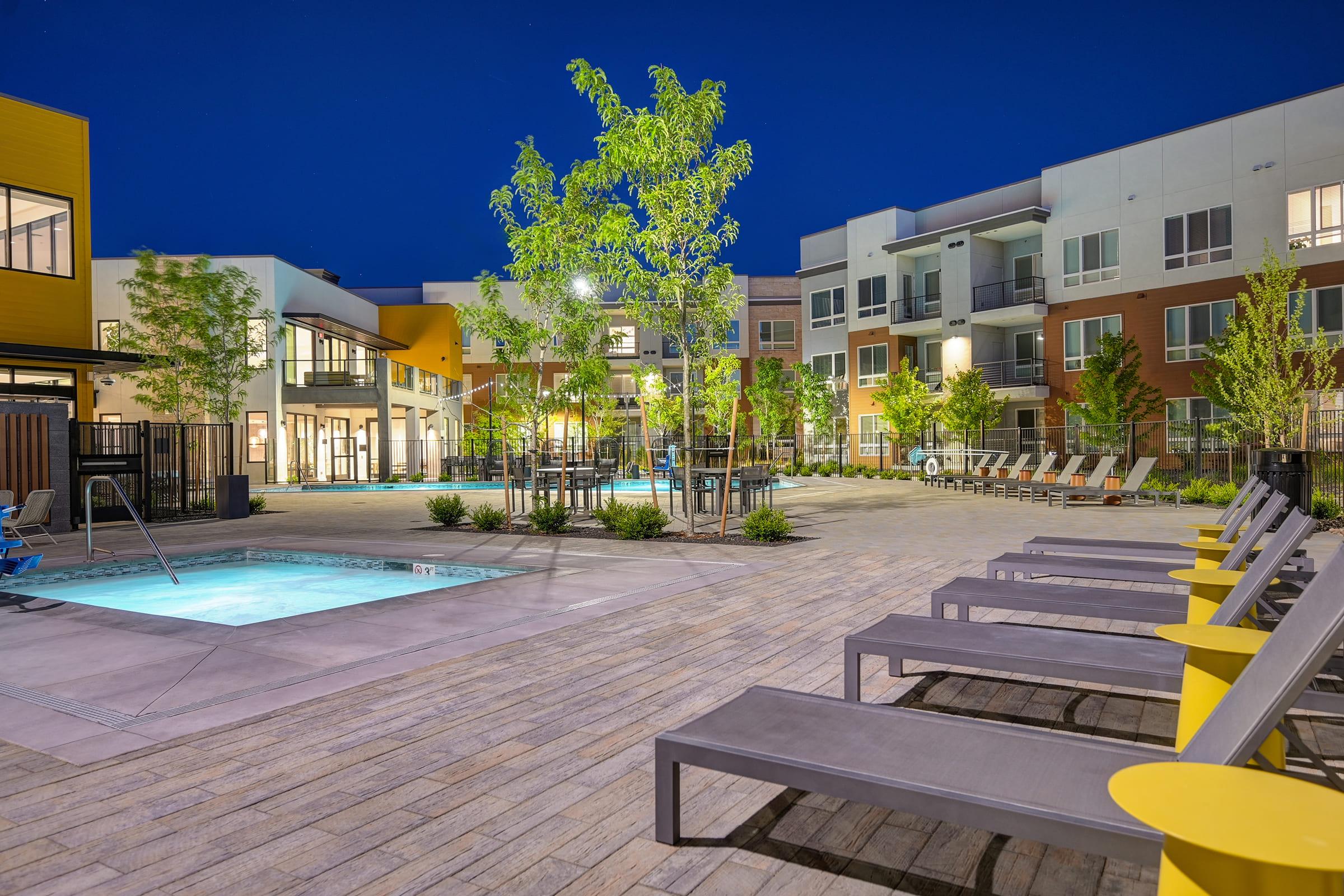 A modern apartment complex courtyard at night featuring a swimming pool, lounge chairs, and landscaped greenery. The buildings have a mix of colors, showcasing contemporary architecture with bright lighting illuminating the area, creating a welcoming atmosphere.