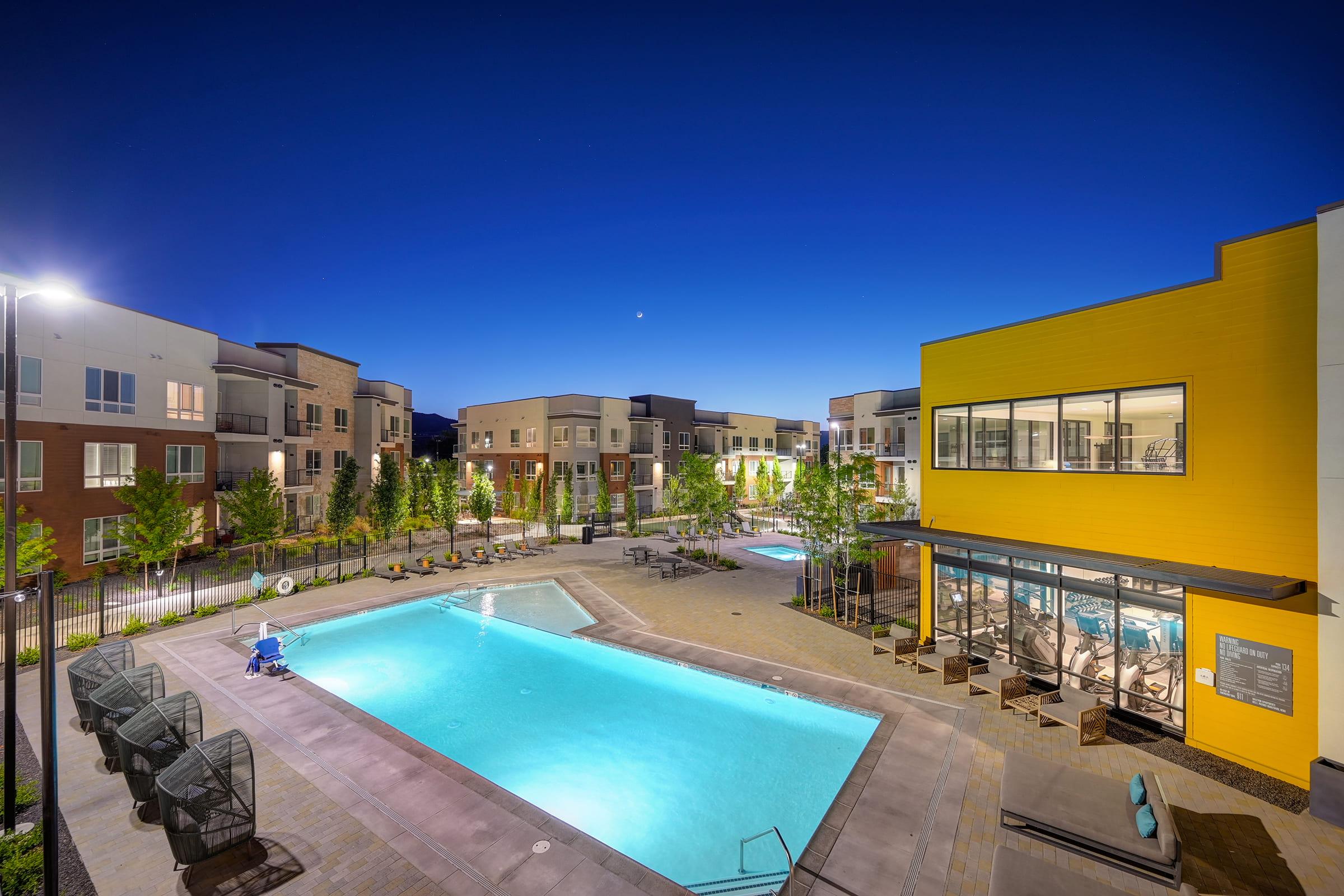 A well-lit apartment complex at night featuring a clear blue sky. The scene includes a large swimming pool surrounded by lounge chairs, landscaped greenery, and modern buildings. A fitness center is visible in the foreground, with illuminated areas enhancing the inviting atmosphere.
