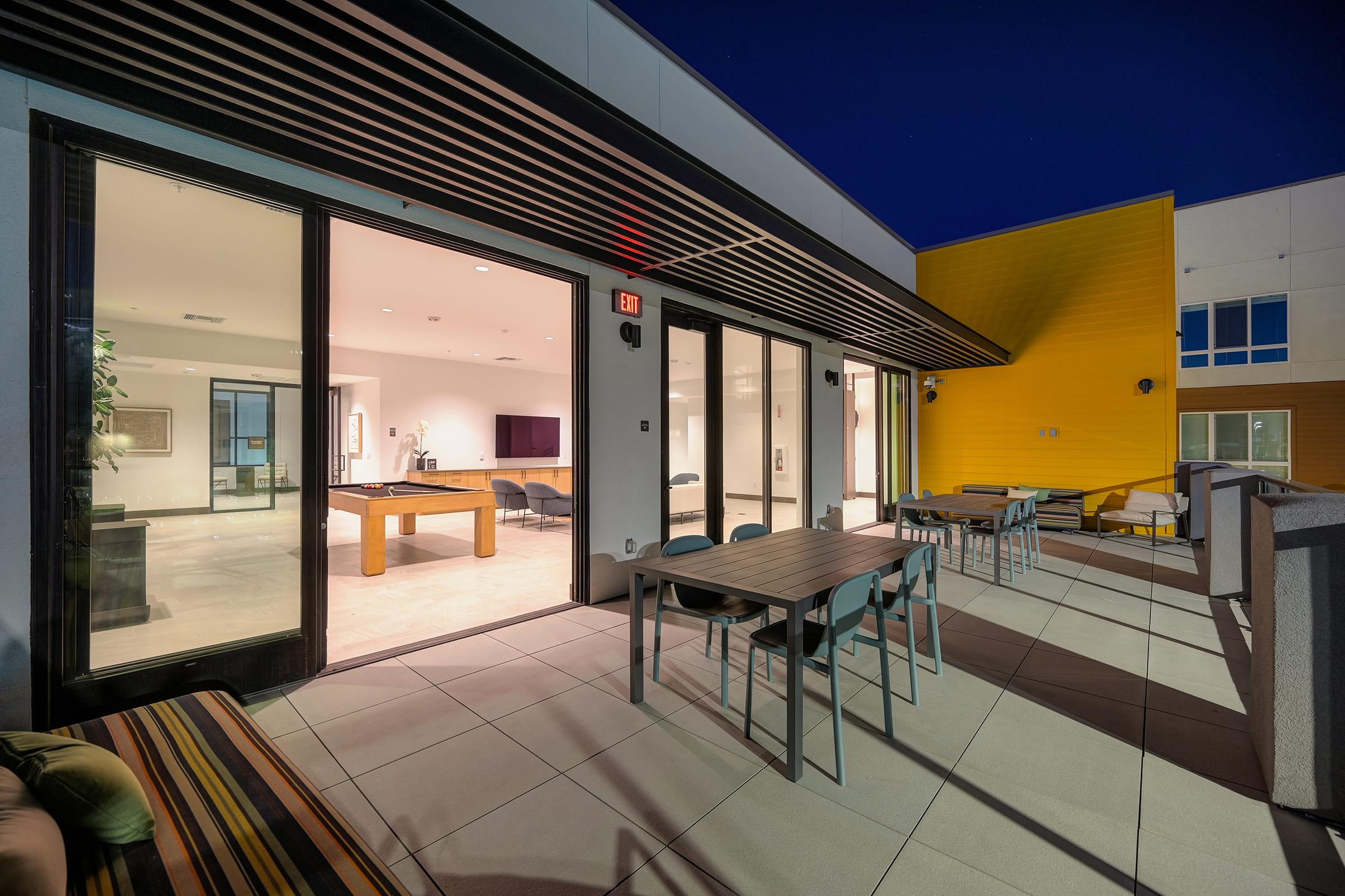 A modern rooftop terrace featuring outdoor furniture, tables, and chairs, with large glass doors leading to an indoor space illuminated at night. The background includes a sleek building exterior and a vibrant yellow wall.