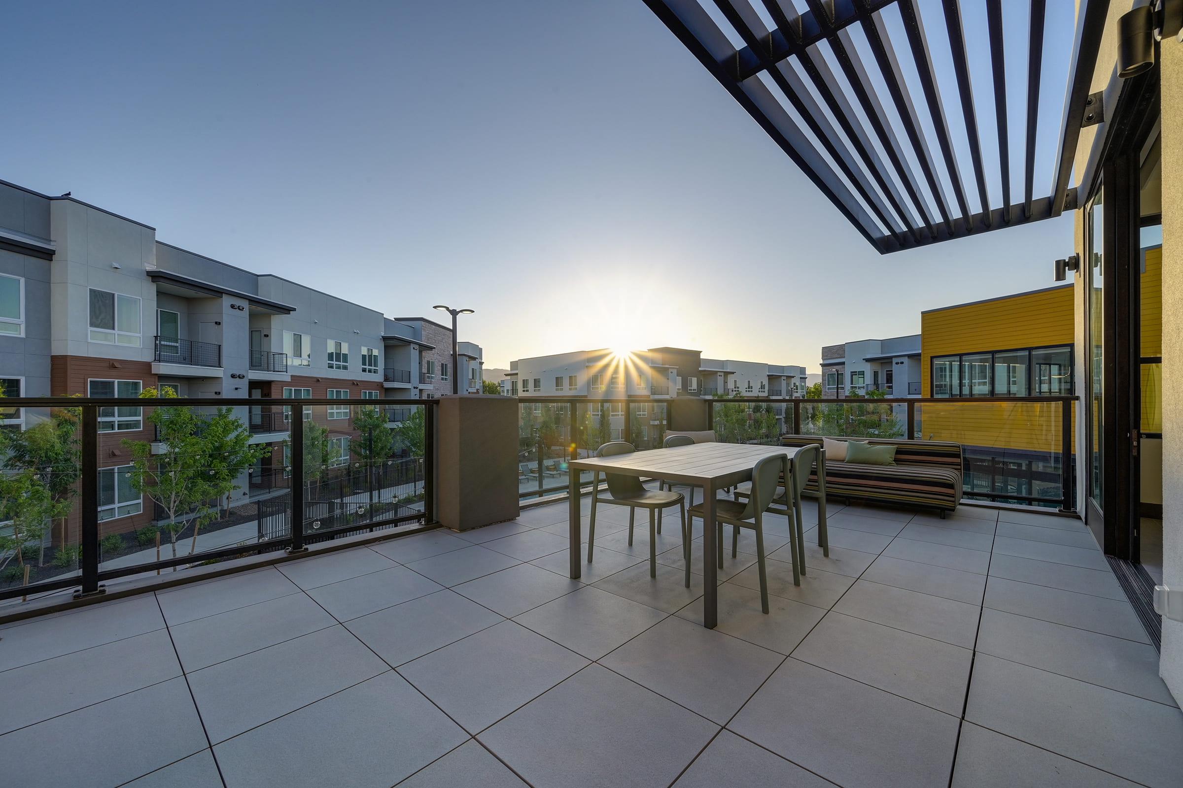 A modern balcony with a dining table and chairs overlooks an apartment complex at sunrise. The scene features sleek architecture, greenery, and a clear sky, creating a serene outdoor space for relaxation and dining.