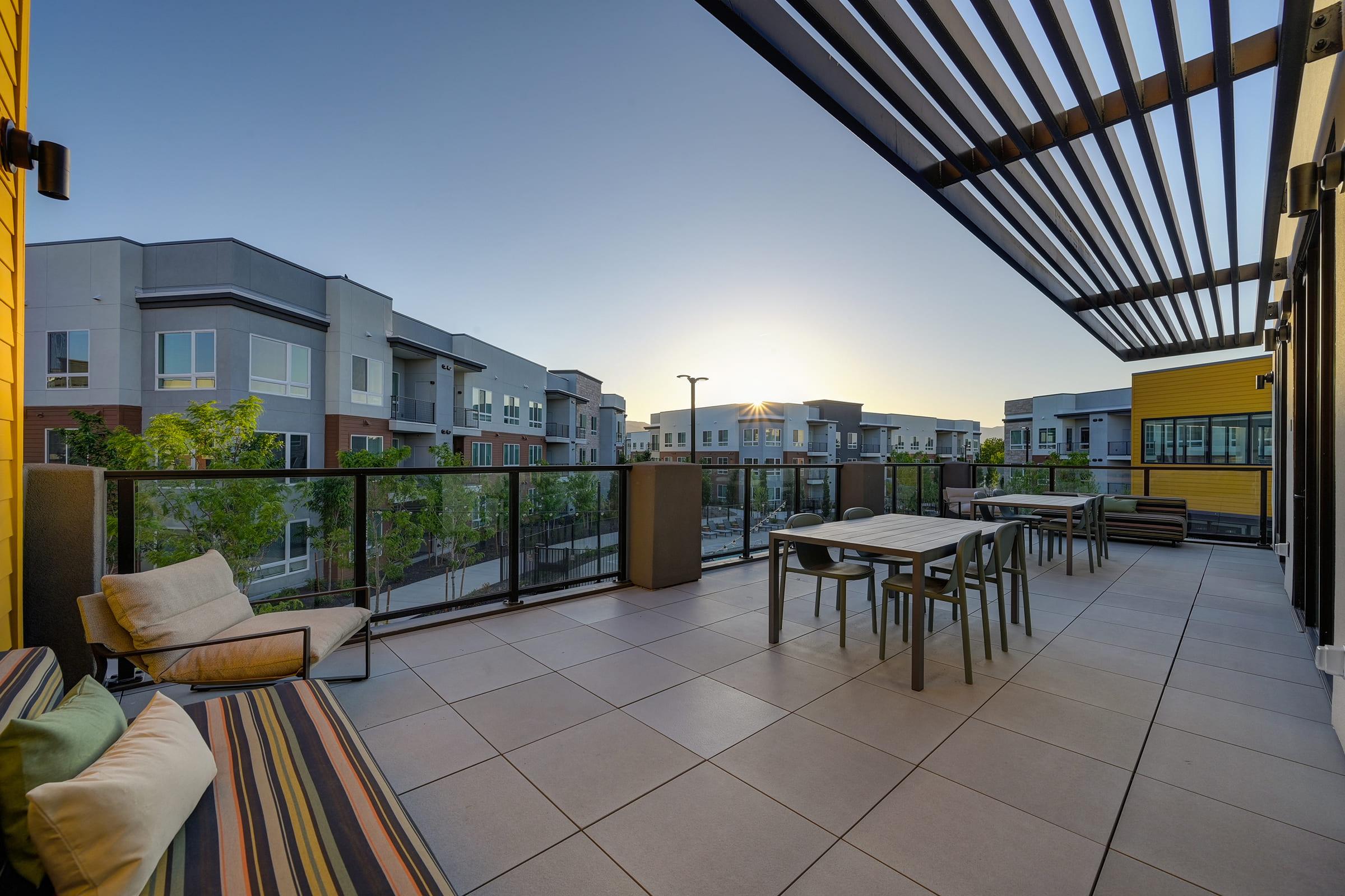 A modern balcony with outdoor seating, featuring striped cushions and a dining table. The background showcases contemporary apartment buildings under a clear sky at sunset. The space is well-lit and inviting, surrounded by greenery, creating a relaxed atmosphere for gatherings or quiet moments.