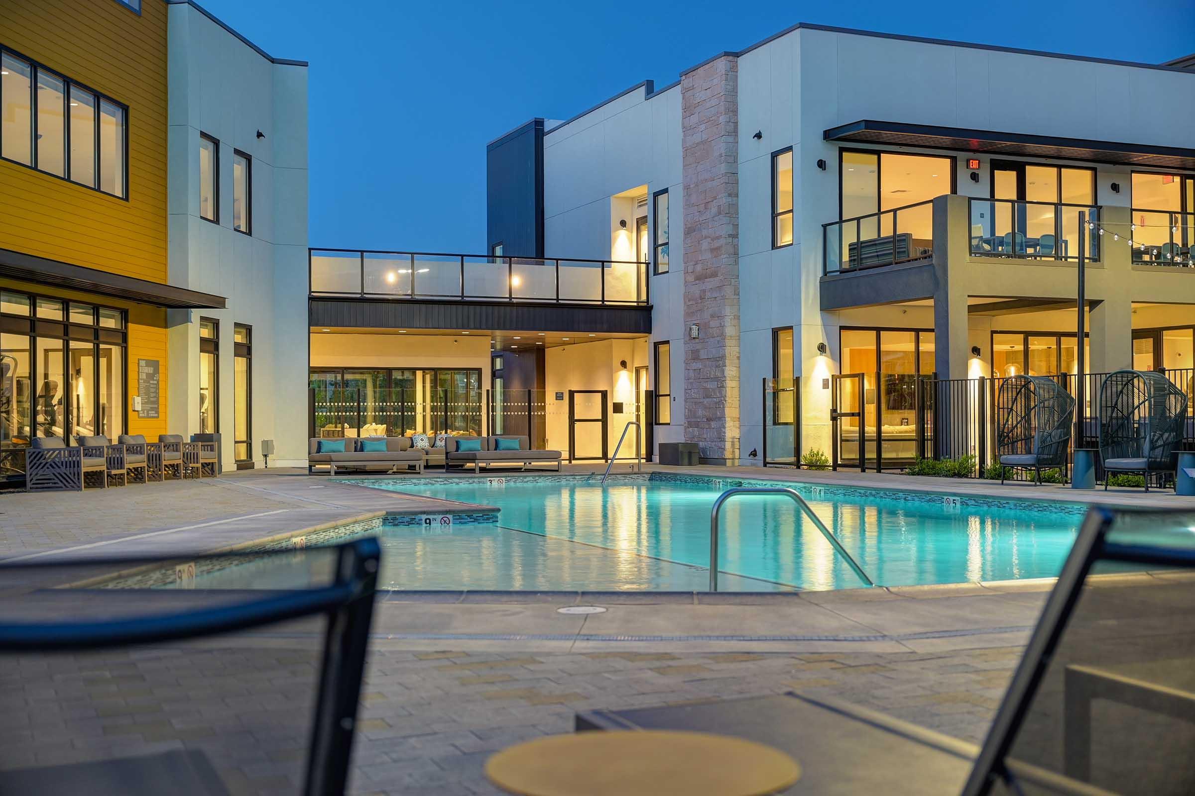 Modern residential complex at dusk featuring a tranquil outdoor pool surrounded by contemporary buildings. Lounge chairs line the pool area, and soft lighting illuminates the scene, creating a welcoming atmosphere for relaxation and community gatherings.