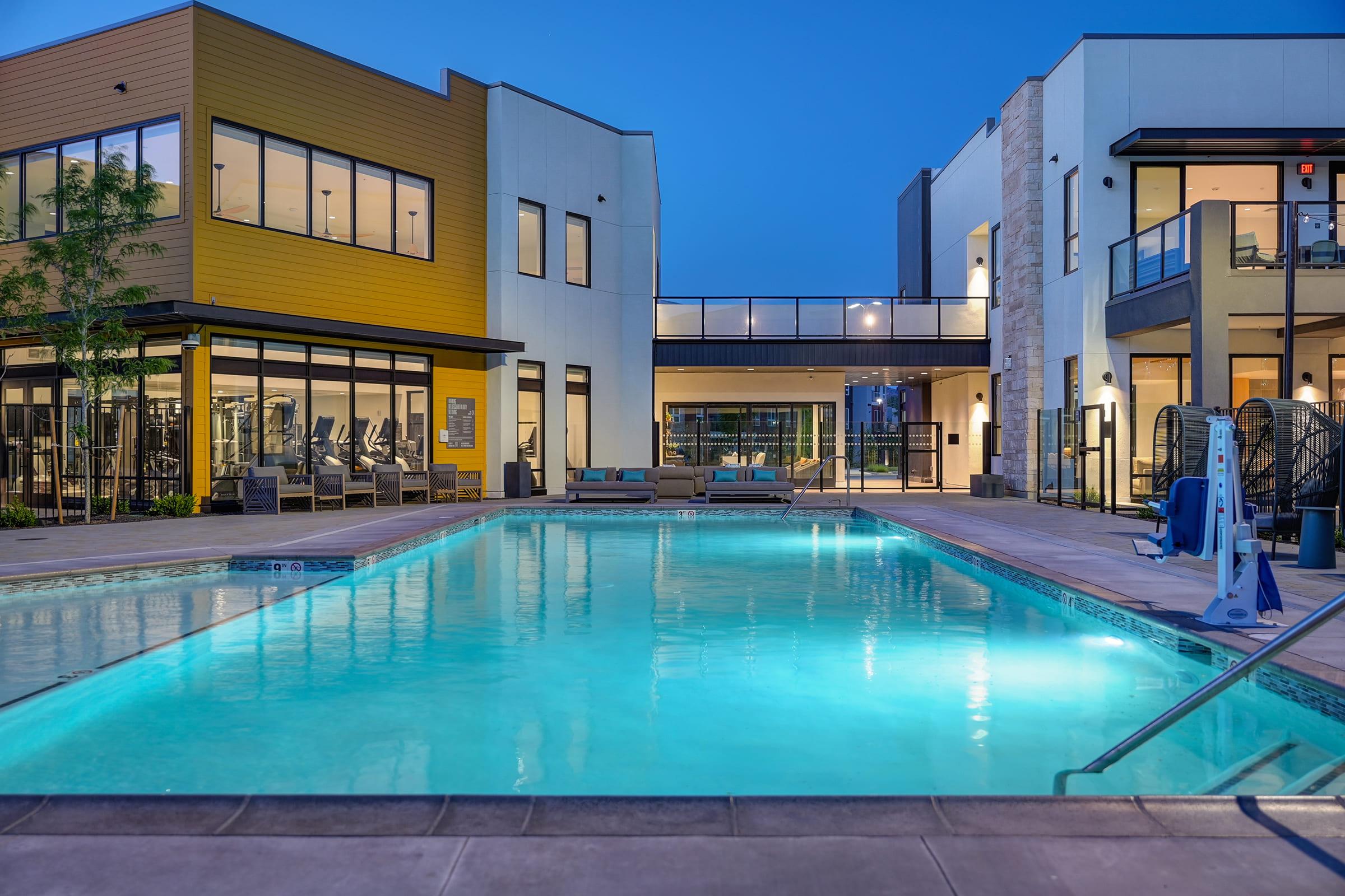 A tranquil evening scene of a modern apartment complex featuring a clear swimming pool surrounded by lounge chairs. The pool is illuminated, reflecting the blue twilight sky, while fitness and recreational areas are visible in the background, highlighting a contemporary architectural design.