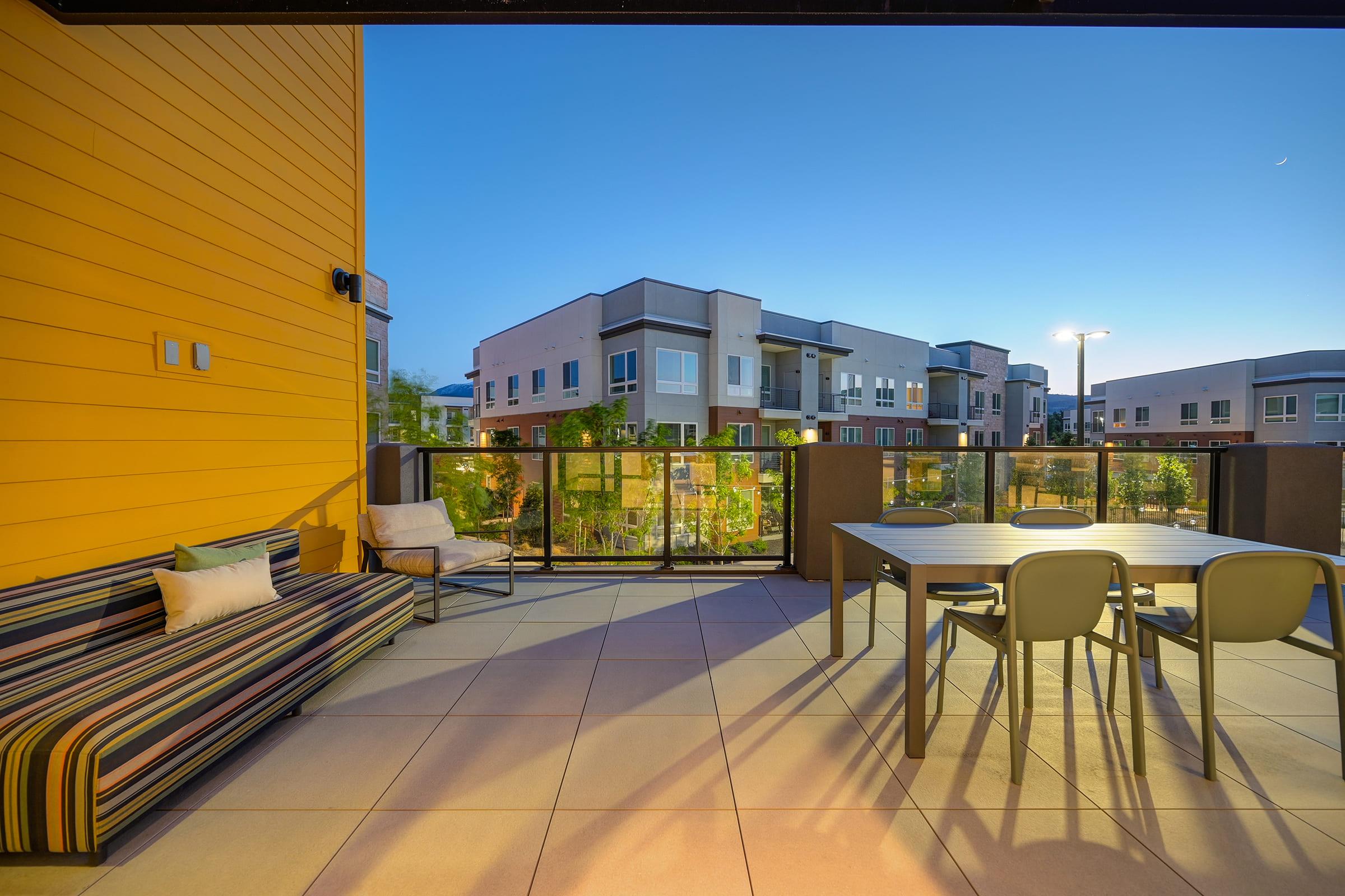 A modern balcony with a striped sofa and a dining table set for outdoor use. The scene showcases an evening sky with a subtle blue hue, overlooking contemporary apartment buildings and landscaped greenery. Soft lighting illuminates the area, creating a cozy atmosphere.