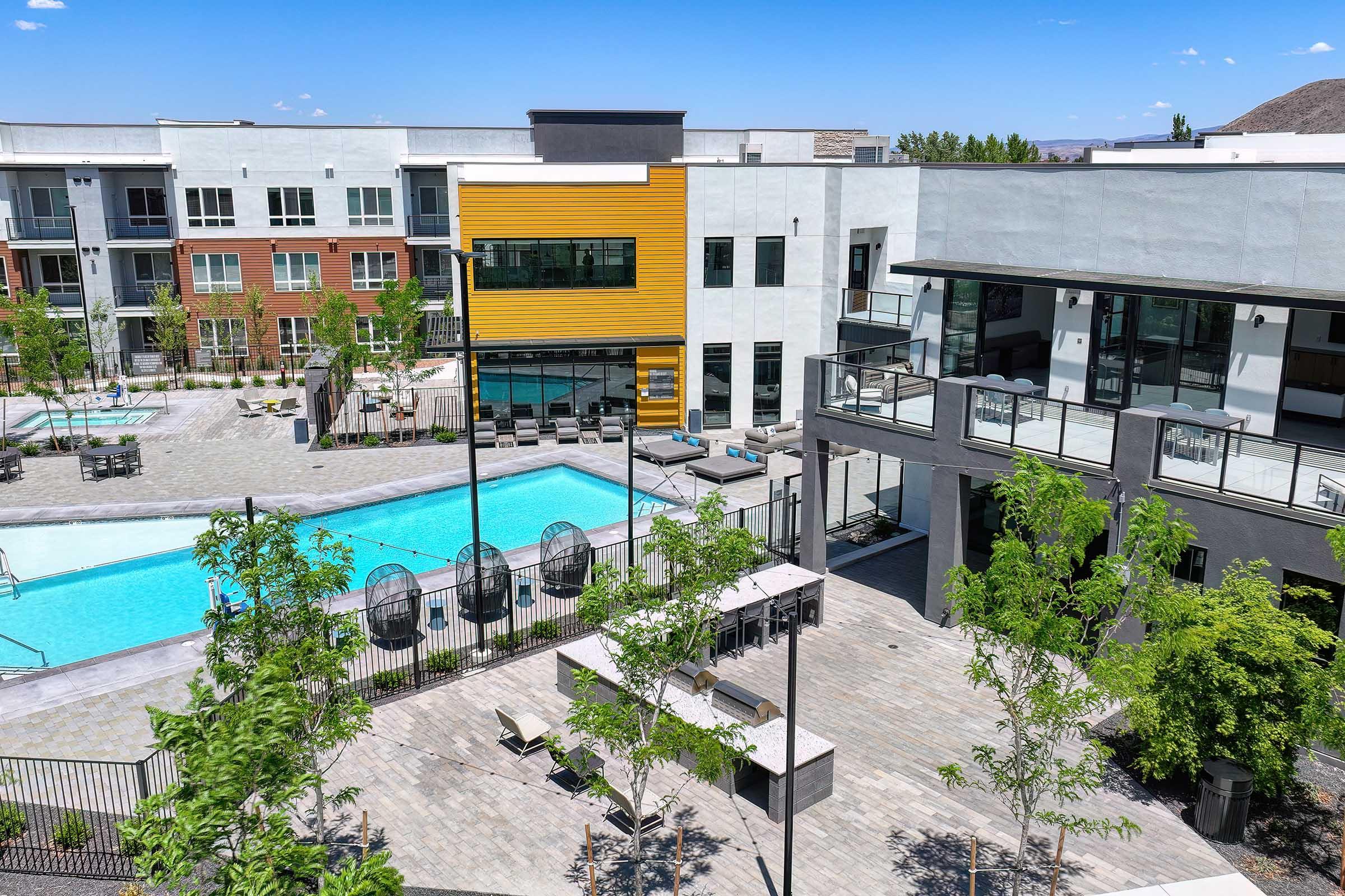 A modern apartment complex with a pool area featuring lounge chairs, surrounded by trees and landscaped paths. The buildings display a mix of colors including yellow and gray, under a clear blue sky.