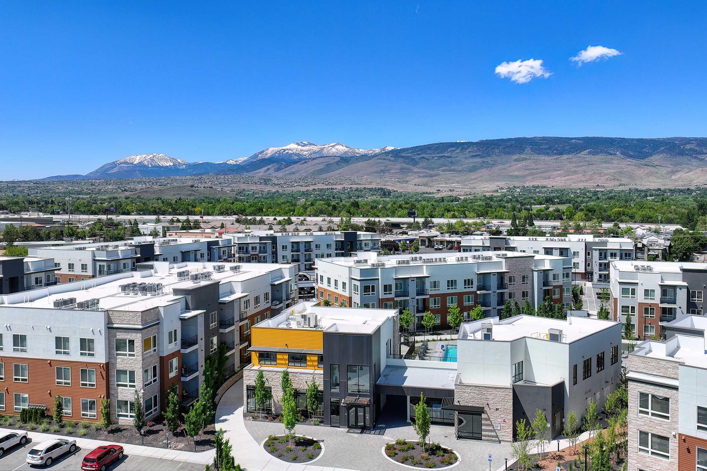 An aerial view of a modern apartment complex surrounded by lush greenery and open spaces, with snow-capped mountains in the background under a clear blue sky. The complex features multiple buildings, a central courtyard, and a swimming pool area.