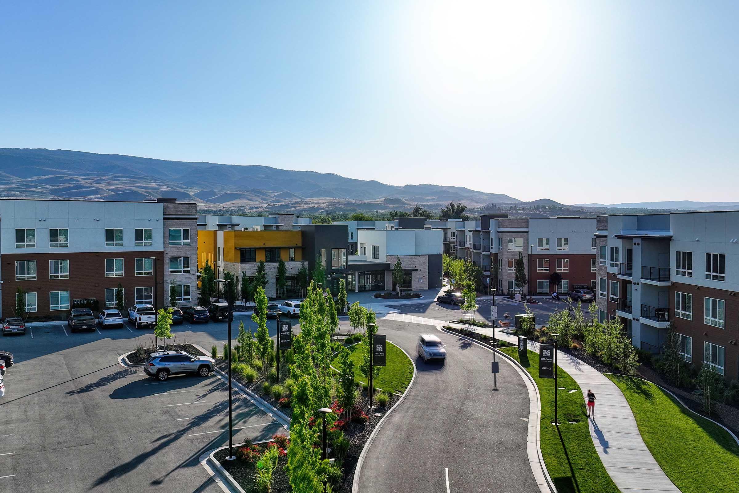 Aerial view of a modern apartment complex with several multi-story buildings in various colors. The area features well-maintained landscaping with greenery and trees, parked cars, and a curving road. In the background, rolling hills and clear blue skies are visible under bright sunlight.