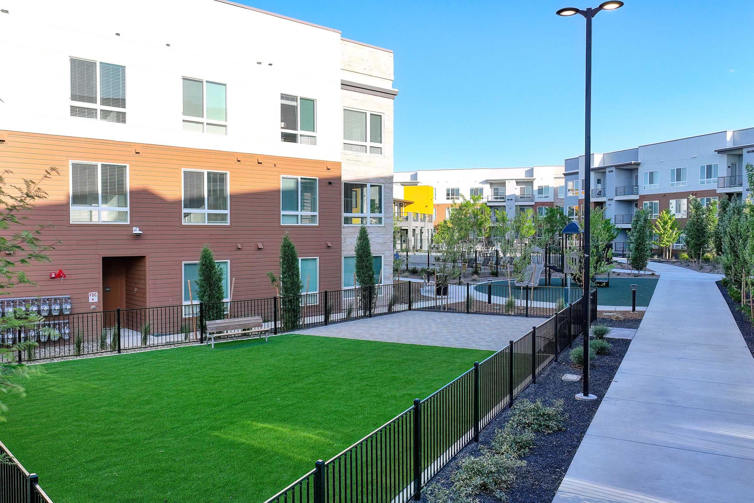 View of a modern apartment complex featuring well-maintained green spaces and pathways. The image shows a grassy area surrounded by trees and a fence, with pathways leading to residential buildings. Nearby, there is a swimming pool area. The setting is bright and welcoming, suggesting a community-oriented environment.