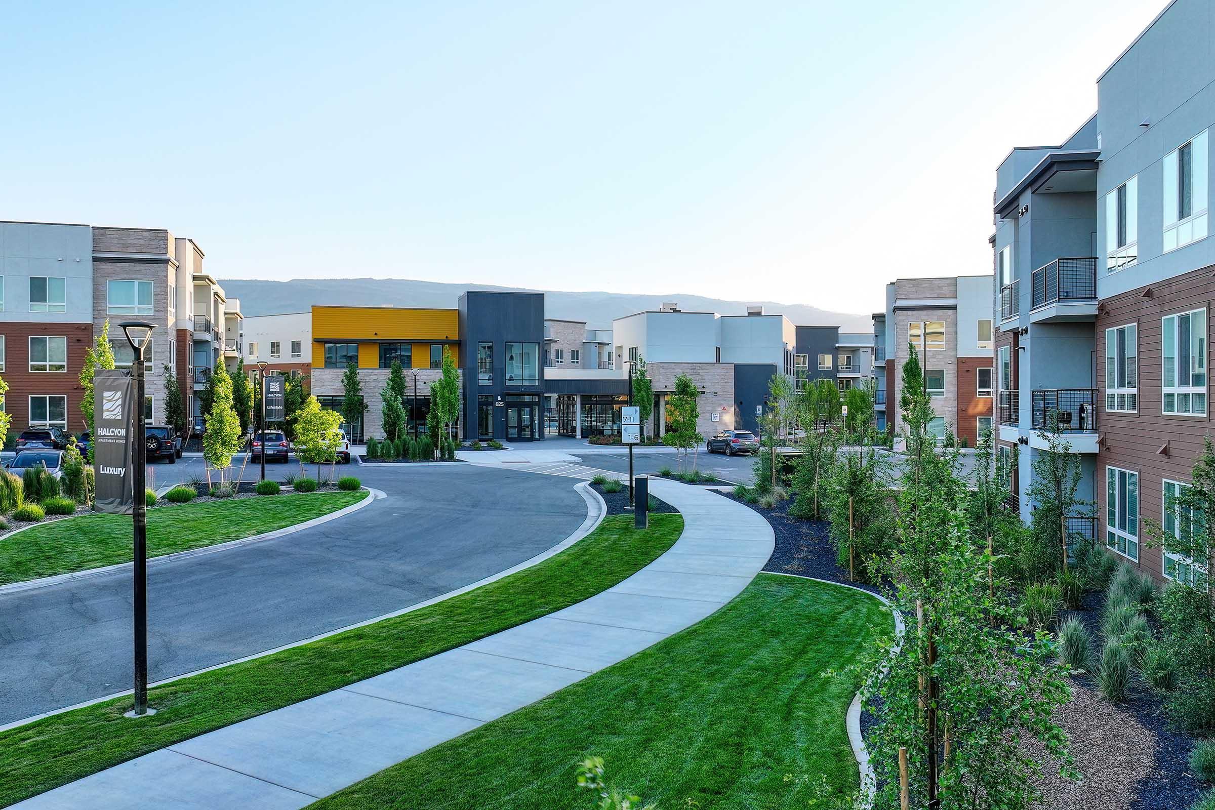 A modern residential complex featuring landscaped grounds, a circular driveway, and contemporary architecture. The image showcases a tranquil environment with green lawns, trees, and nearby parking, set against a backdrop of mountains and clear skies.