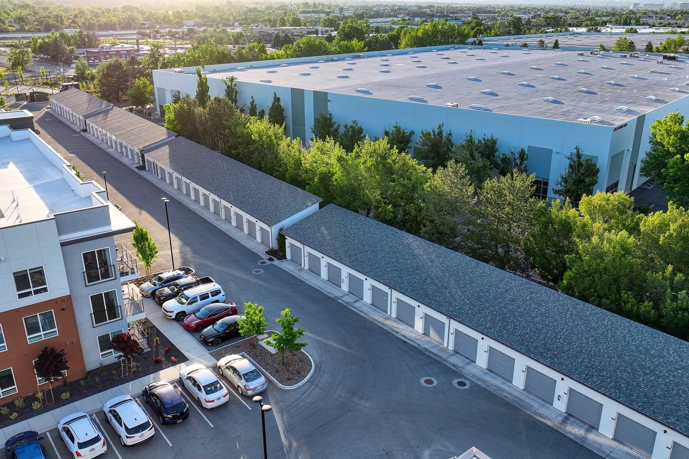 Aerial view of a modern storage facility with several storage units lined along a paved road, surrounded by trees. Nearby, a multi-story residential building and a parking lot with various cars are visible. The scene is set in a suburban area with a large warehouse in the background.