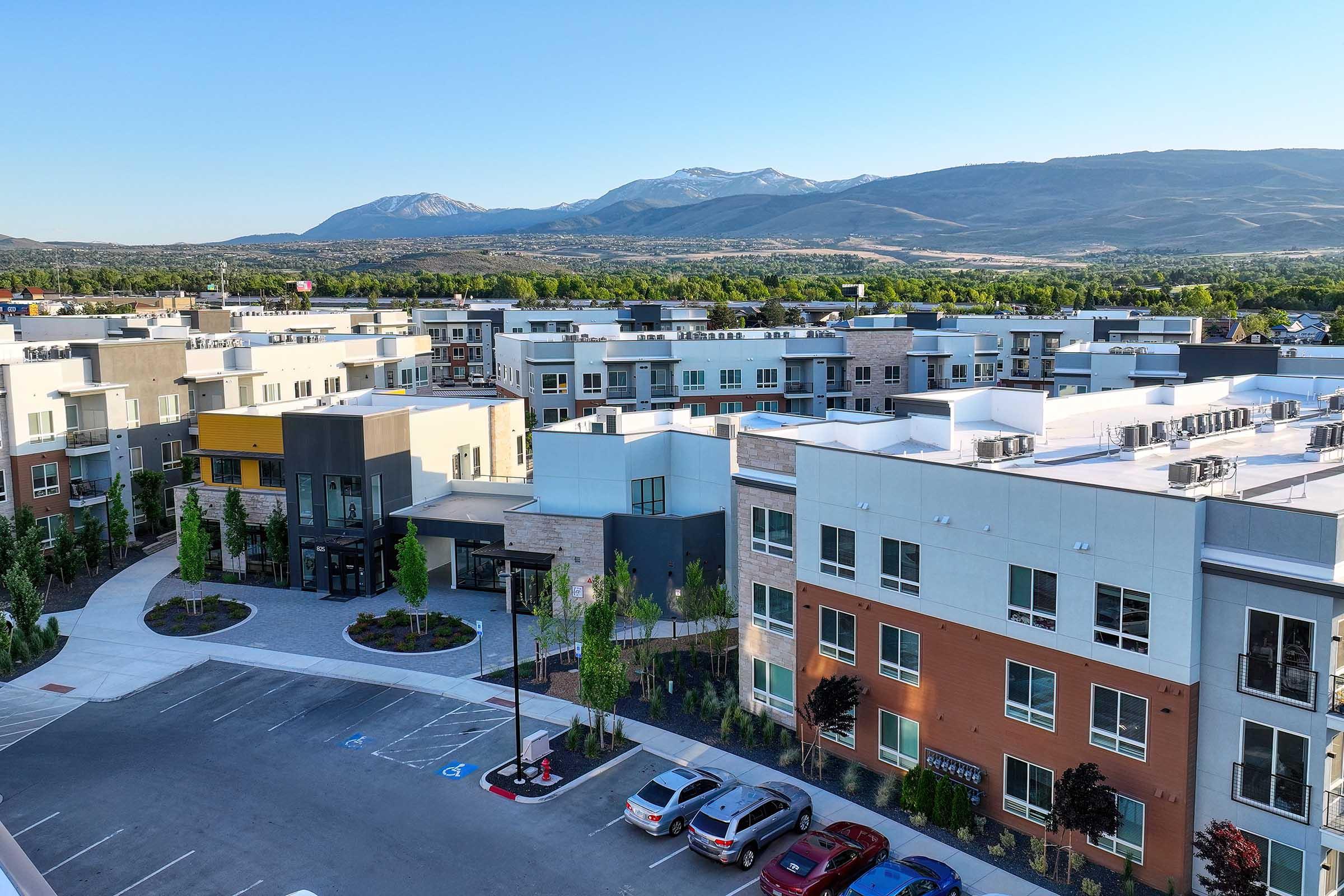 A scenic view of a modern residential complex with multiple buildings, surrounded by landscaped greenery. In the background, mountains rise against a clear blue sky, and a parking area is visible in the foreground, showcasing well-maintained architecture and pleasant outdoor spaces.