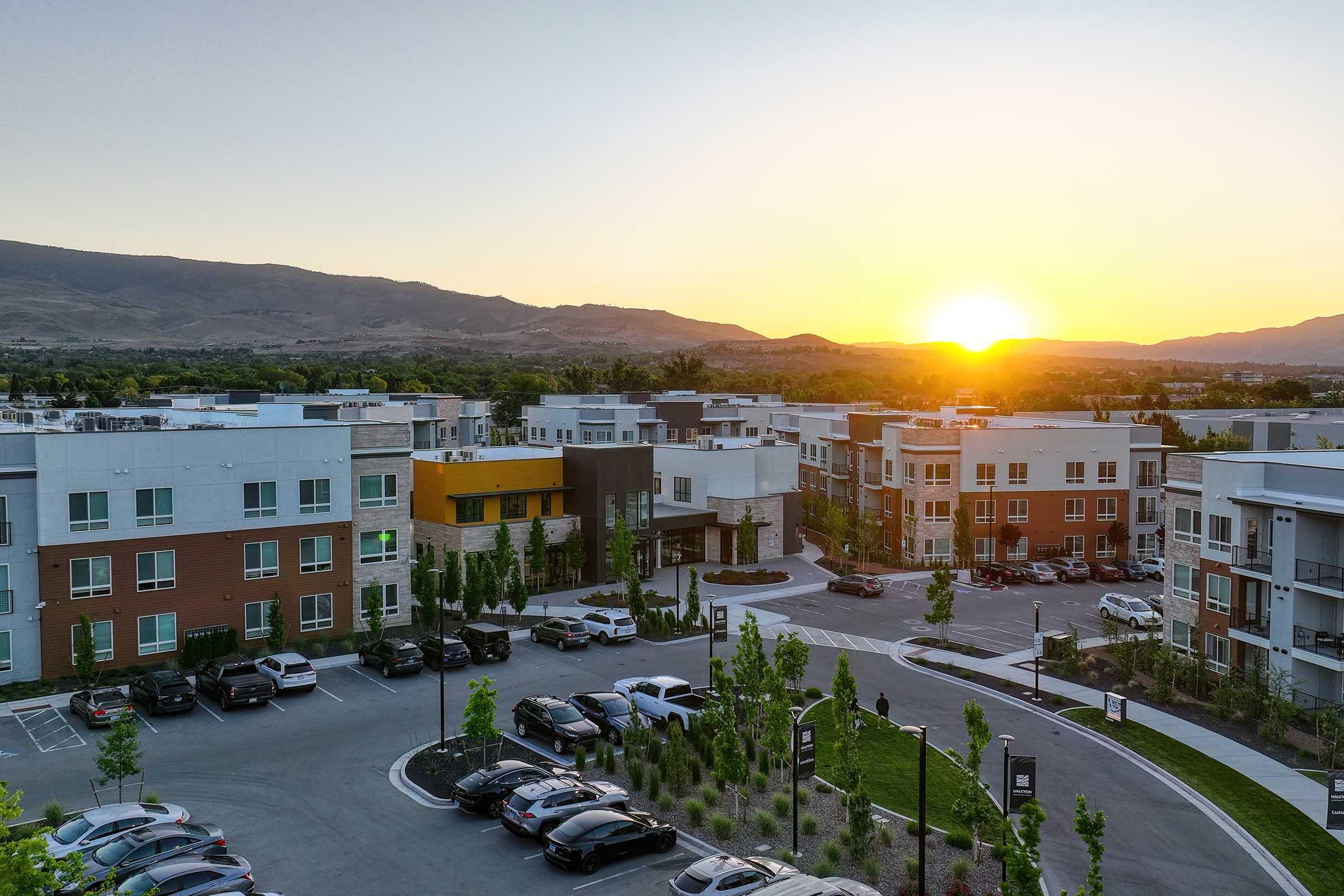 A scenic view of modern apartment buildings nestled in a residential area at sunset. The sun is setting behind distant mountains, casting warm light over the landscape. Cars are parked in a lot, and trees and greenery are visible throughout the area, creating a pleasant atmosphere.