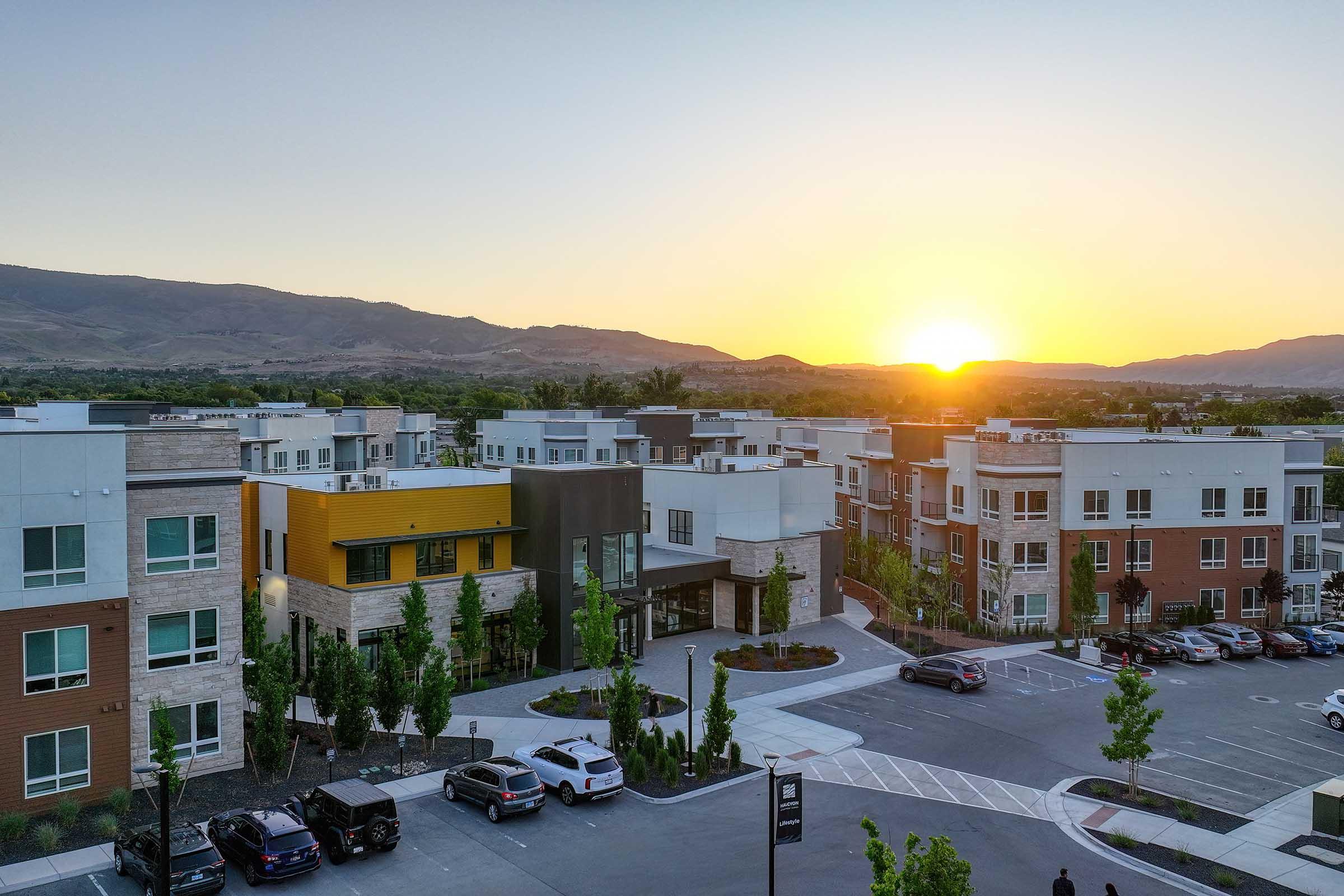 A view of modern apartment buildings in a residential area at sunset, with mountains in the background. The scene features a parking lot with several cars and landscaped green spaces. The warm colors of the setting sun create a picturesque atmosphere.
