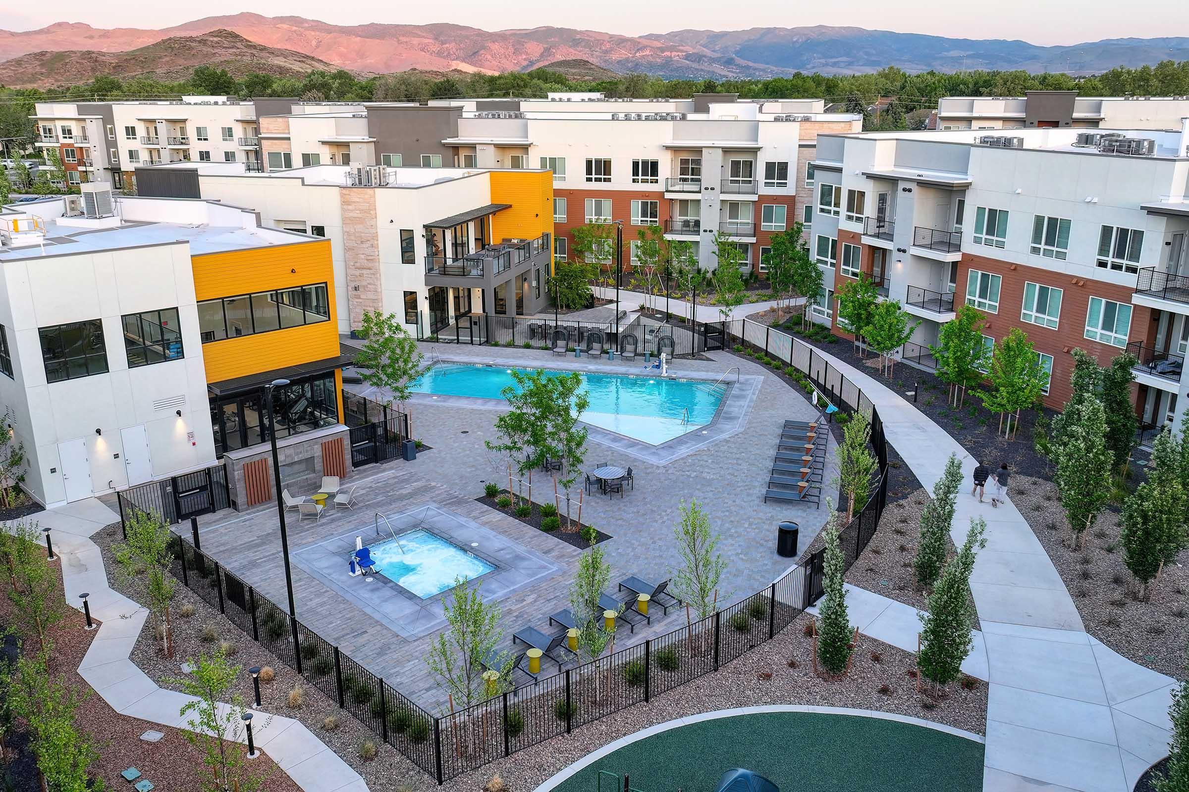 Aerial view of a modern residential complex featuring a swimming pool and hot tub area surrounded by lounge chairs. Green trees and shrubs are planted around the pool, with well-maintained paths and landscaping. In the background, several multi-story buildings can be seen, set against a mountainous scenery.
