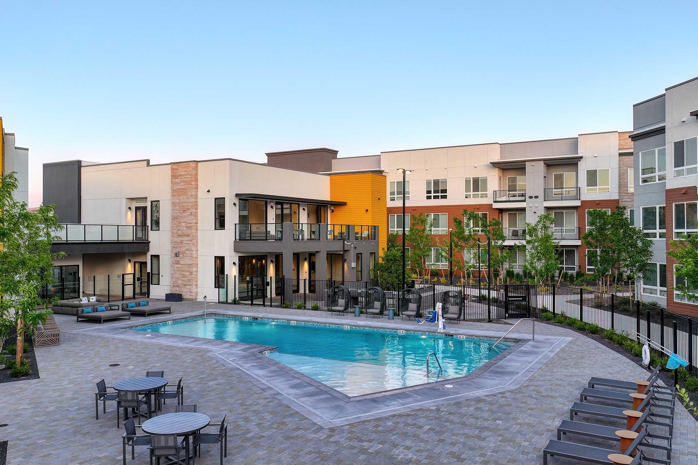 A modern apartment complex with a well-maintained swimming pool surrounded by lounge chairs and tables. Green trees line the pool area, and contemporary buildings with balconies and large windows can be seen in the background against a clear blue sky.