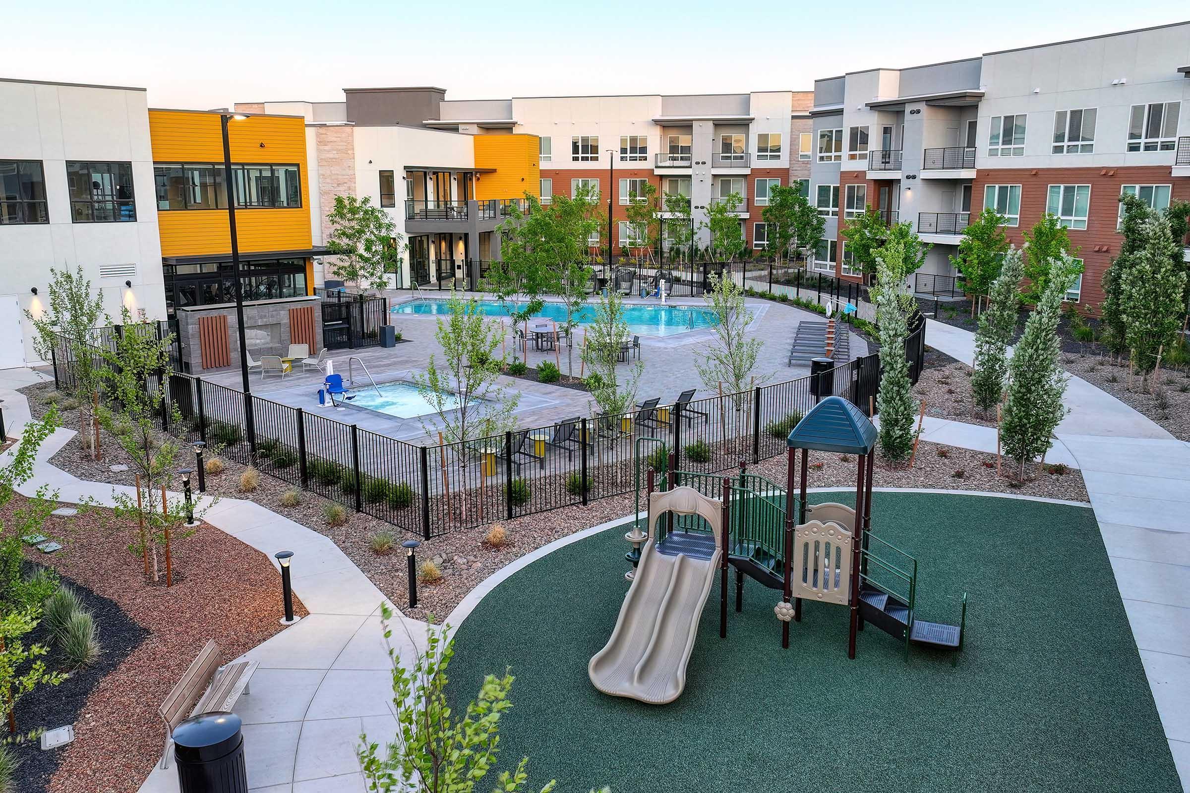 A modern residential courtyard featuring a swimming pool, playground with a slide, and landscaped greenery. The area is enclosed by a black fence, with several apartment buildings surrounding the space, showcasing a blend of contemporary architecture and outdoor amenities.