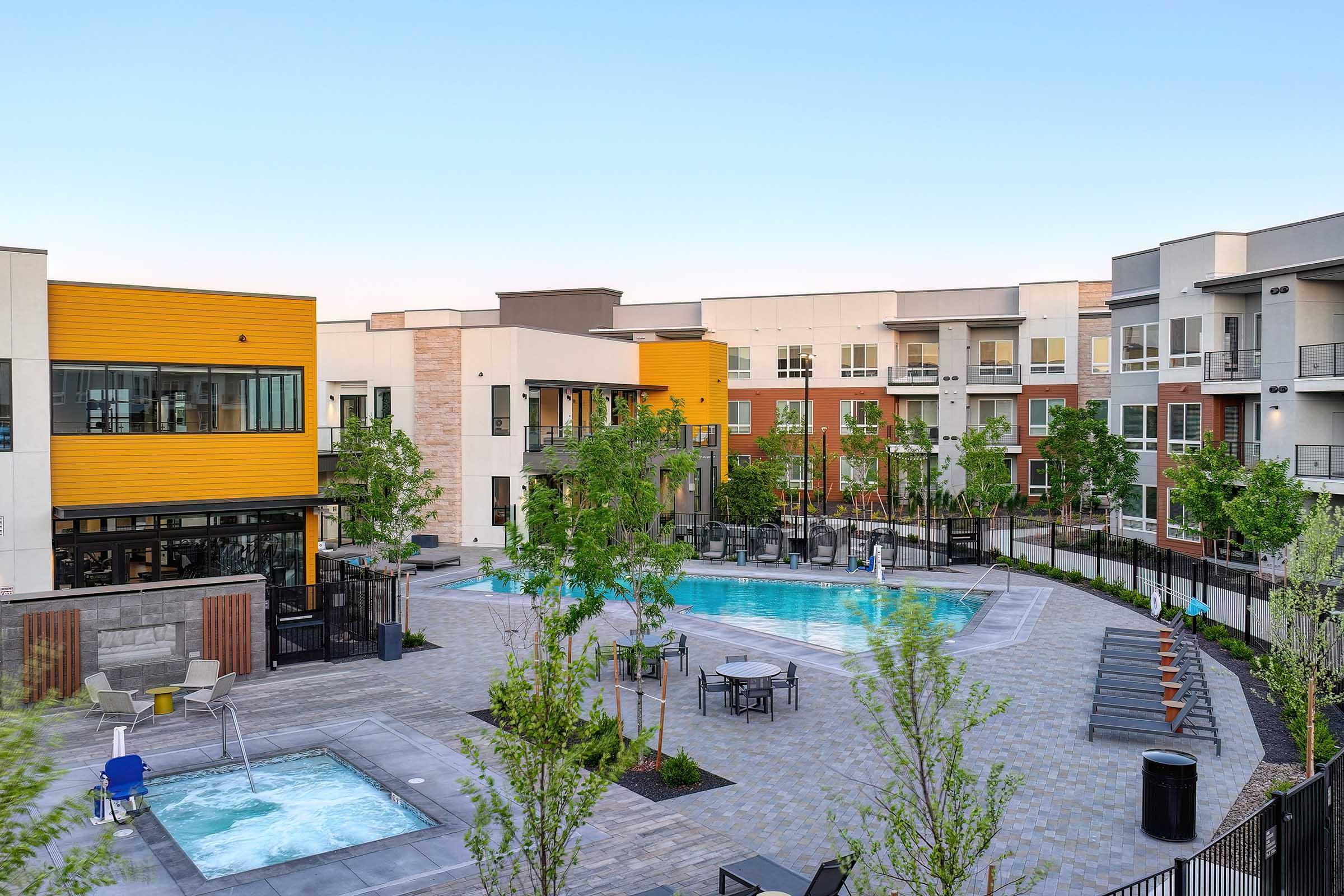 A modern apartment complex featuring a central courtyard with two swimming pools, surrounded by landscaped greenery and seating areas. The buildings exhibit a mix of yellow, gray, and red facades, with clear blue skies overhead.