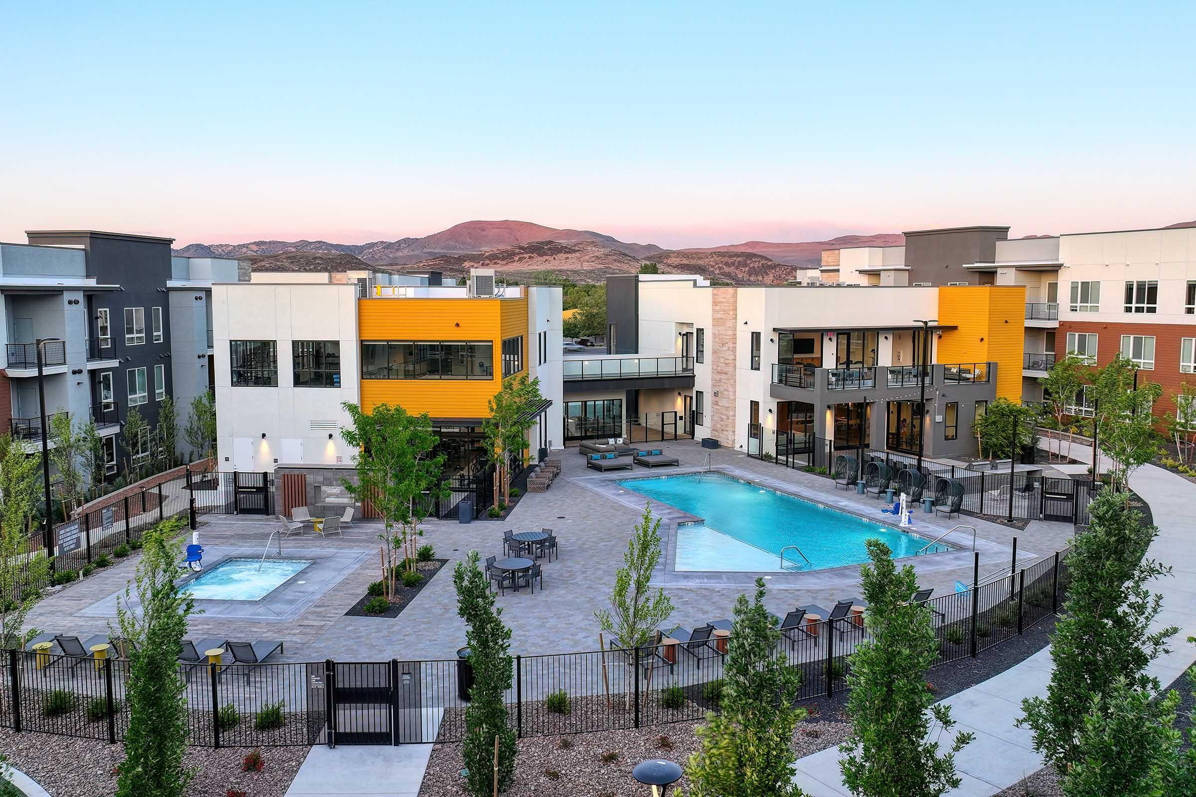 A modern apartment complex featuring a swimming pool and hot tub area surrounded by landscaped greenery. The buildings have a contemporary design with a mix of colors, and there's a clear sky overhead, suggesting a serene environment in a residential area.