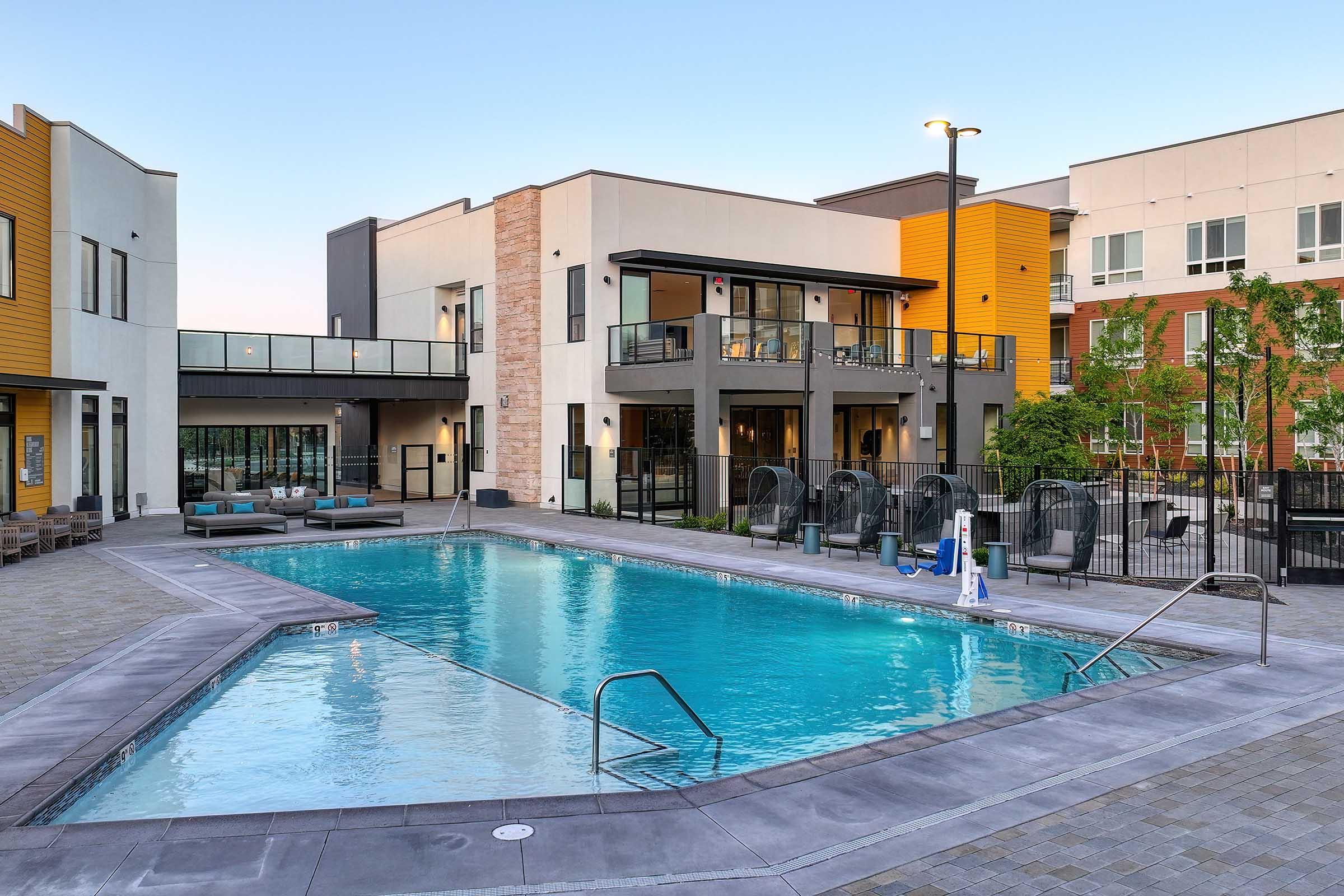 A modern residential complex featuring a sparkling blue swimming pool surrounded by lounge chairs and tables. The buildings are designed with a mix of stone and wood exteriors, showcasing balconies and large windows. Lush greenery adds to the inviting atmosphere, with twilight lighting enhancing the scene.