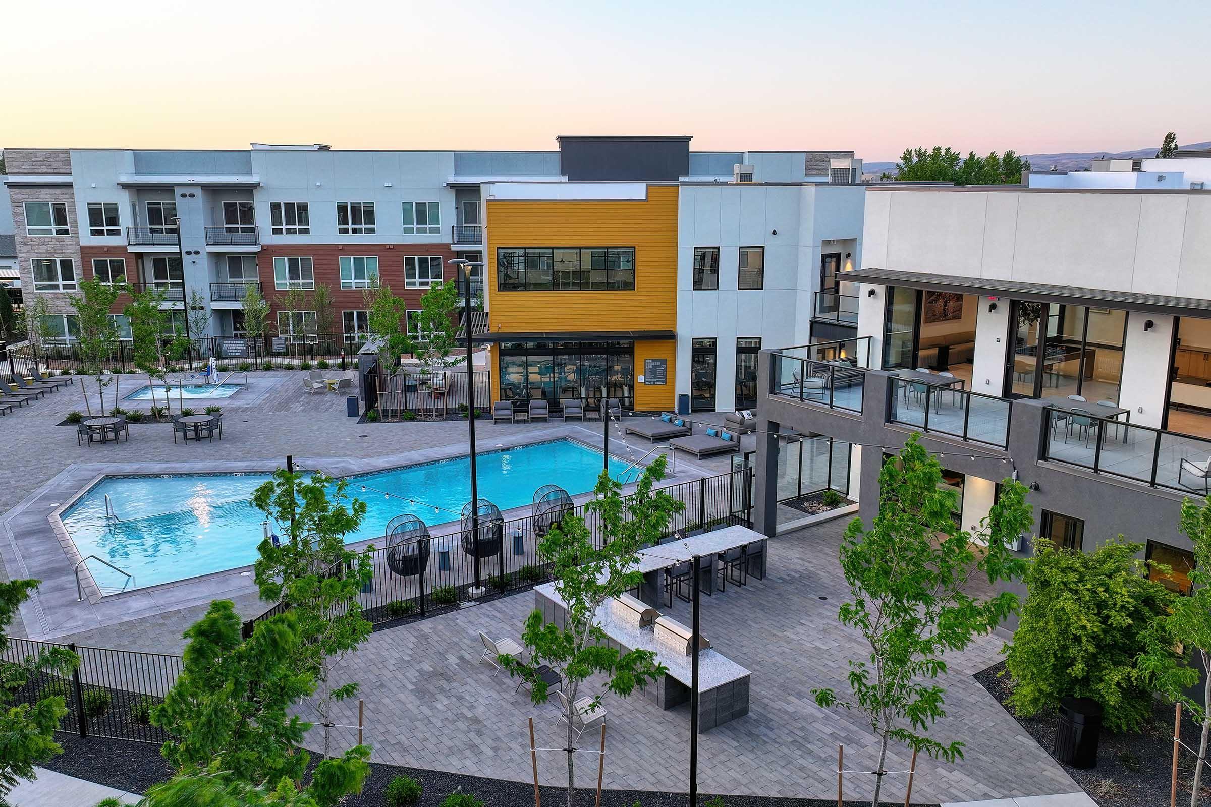 View of a modern apartment complex featuring a landscaped courtyard with a swimming pool. Surrounding the pool are lounge areas with seating, decorative trees, and well-maintained paths. The buildings have a mix of colors, including brick and yellow accents, set against a clear evening sky.