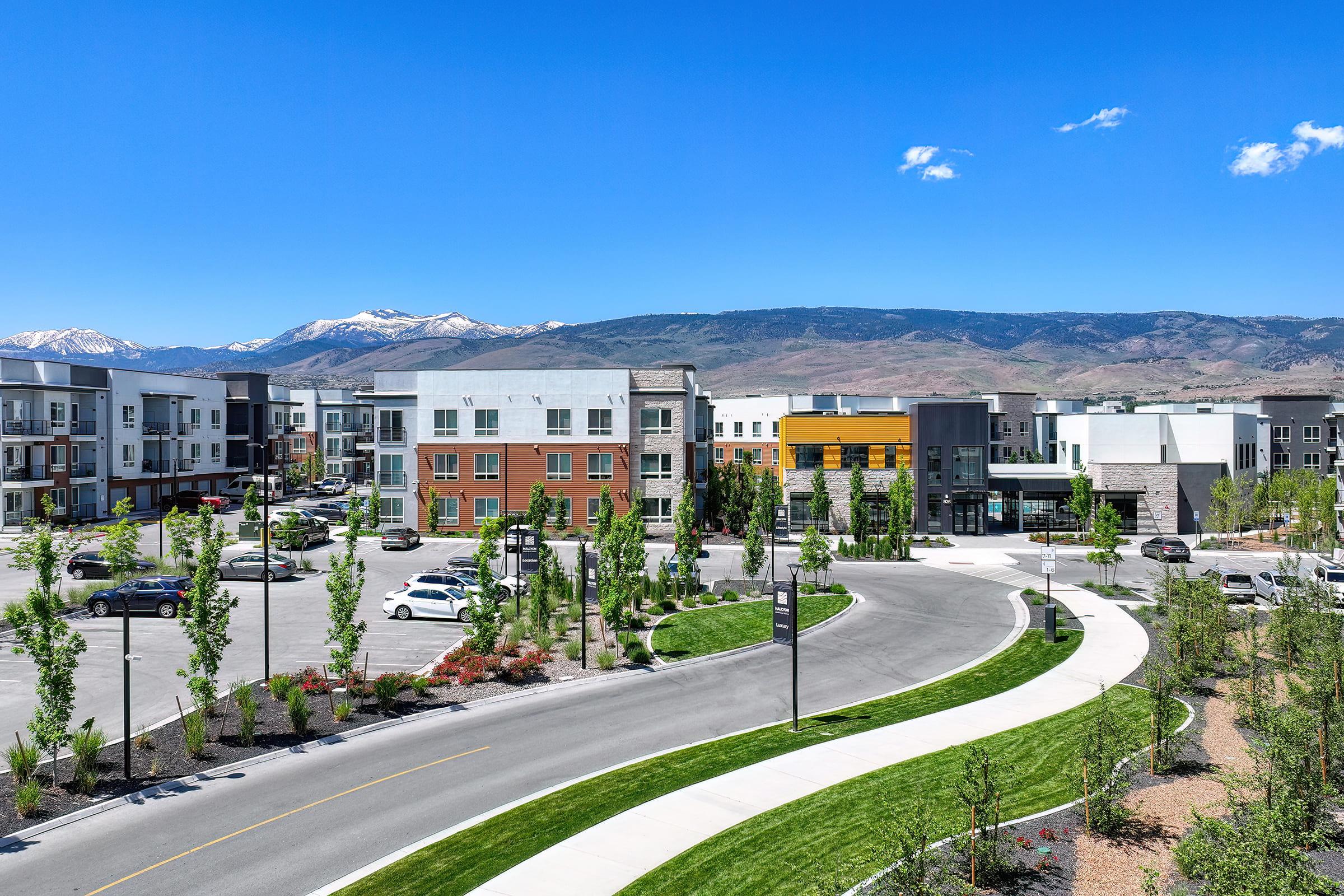 A modern residential complex with several multi-story buildings surrounded by greenery. The foreground features a curved pathway and a parking lot filled with cars. In the background, mountains are visible under a clear blue sky, adding a scenic backdrop to the urban environment.