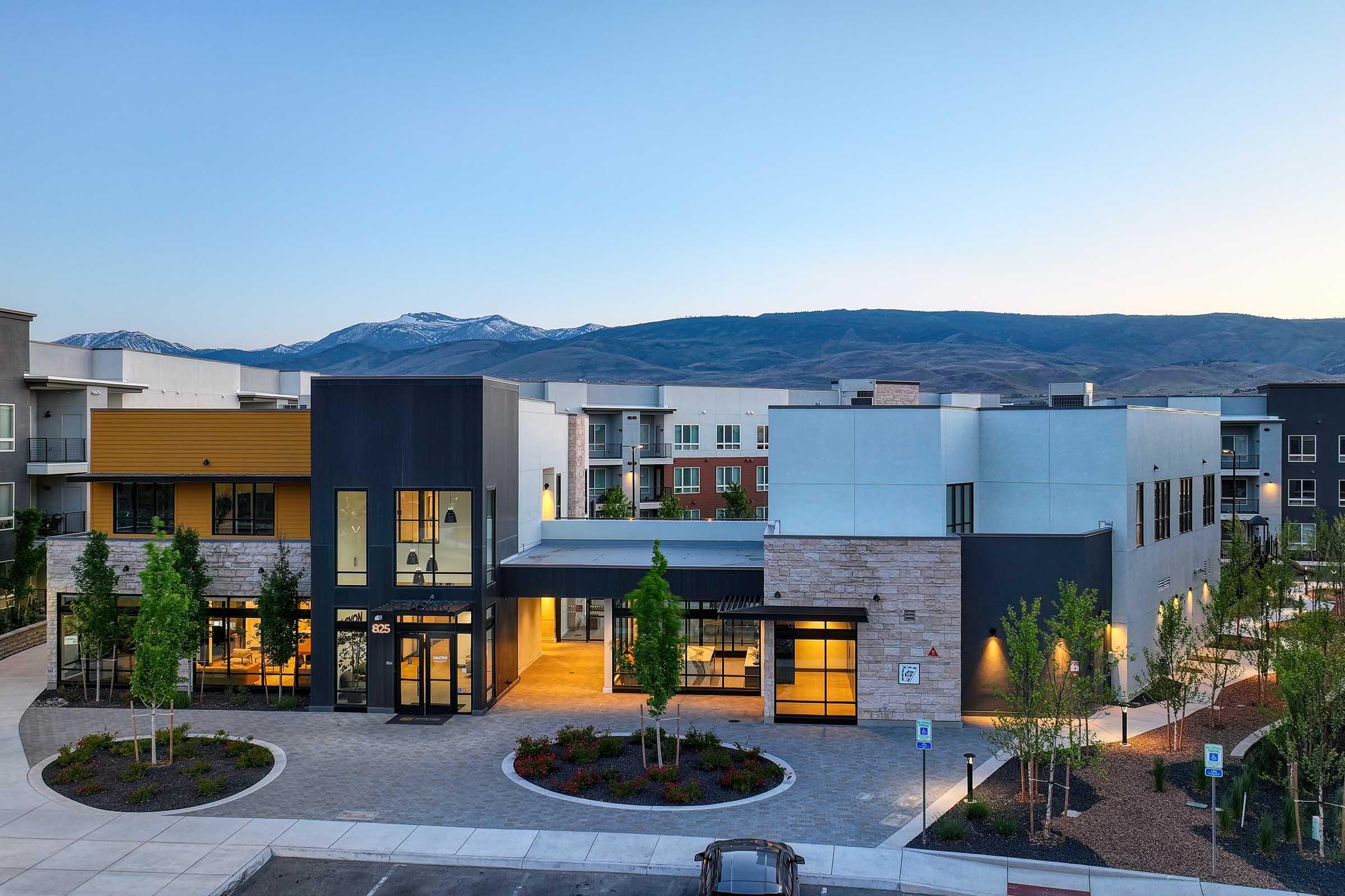 Modern commercial building in a scenic setting with mountains in the background. The structure features a mix of materials, including stone and smooth concrete, surrounded by landscaped greenery and walkways. The sky is clear, indicating early evening light.