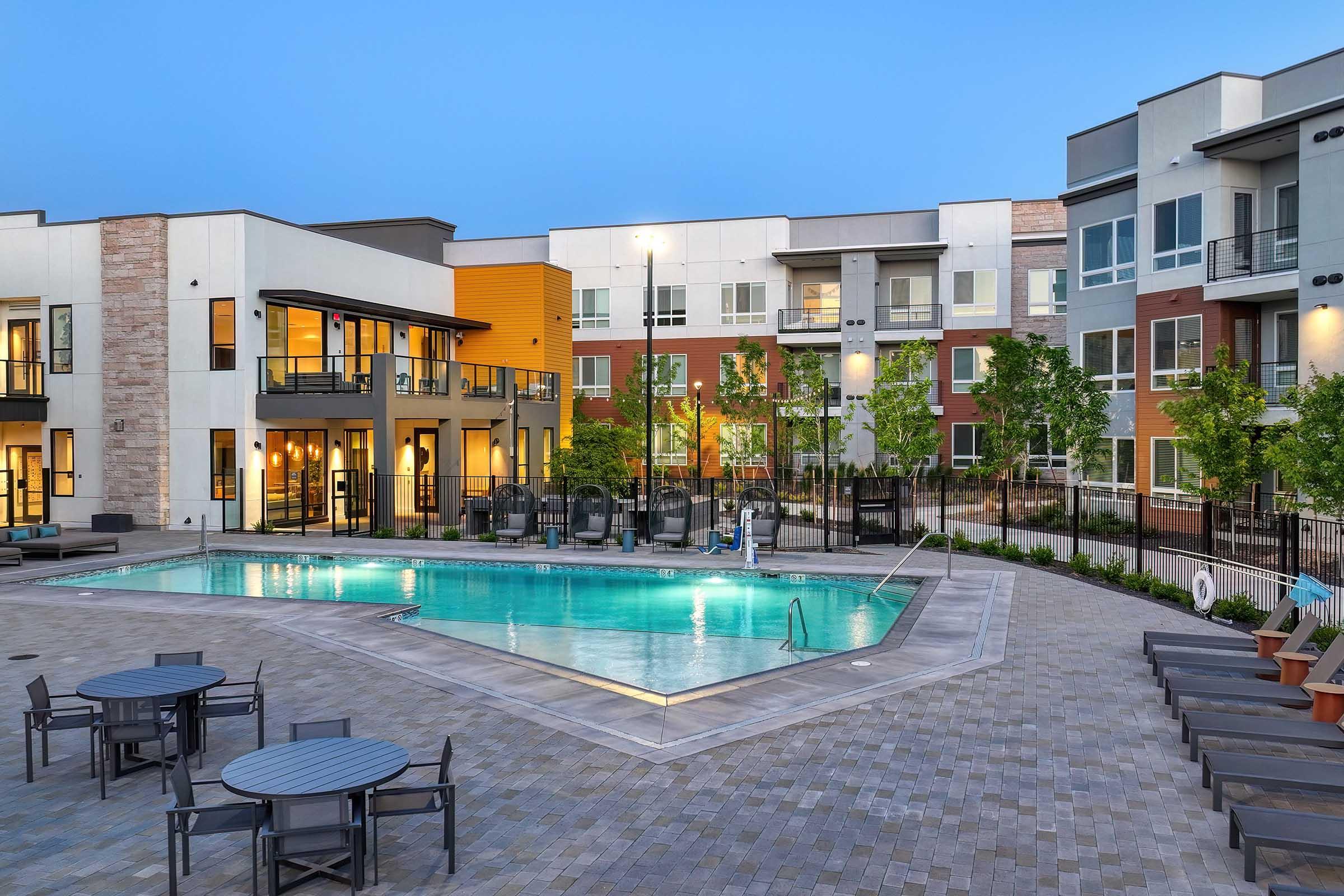 A modern apartment complex courtyard featuring a swimming pool surrounded by lounge chairs and dining tables. The buildings showcase contemporary architecture with large windows and a mix of colors. Trees and landscaping enhance the outdoor space, creating a welcoming atmosphere.