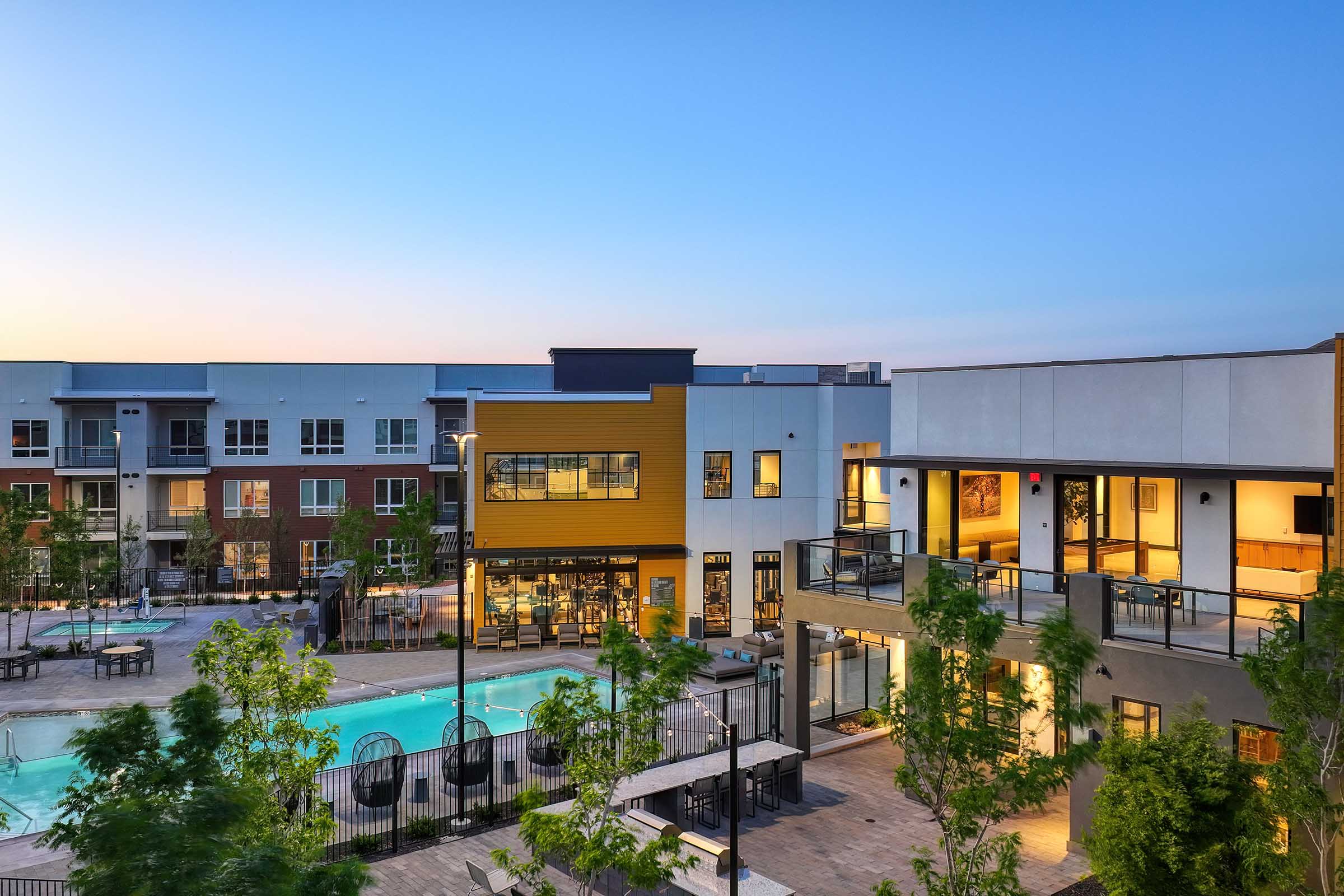 A modern apartment complex with a swimming pool surrounded by lounge chairs and trees. The buildings feature a mix of colors and designs, with balconies and outdoor seating areas. The sky is clear, suggesting early evening or dusk lighting.