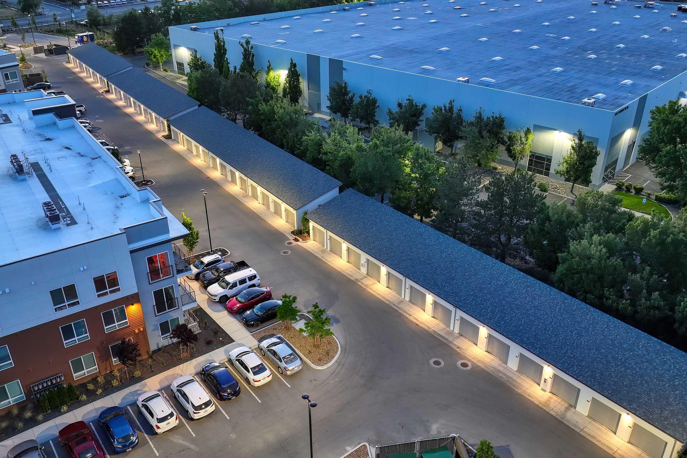 An aerial view of a parking area with multiple cars parked along a curved driveway, surrounded by buildings. On one side, there's a two-story apartment complex with outdoor lighting, and on the other, a large industrial building. Trees and landscaping are visible in the surrounding area.