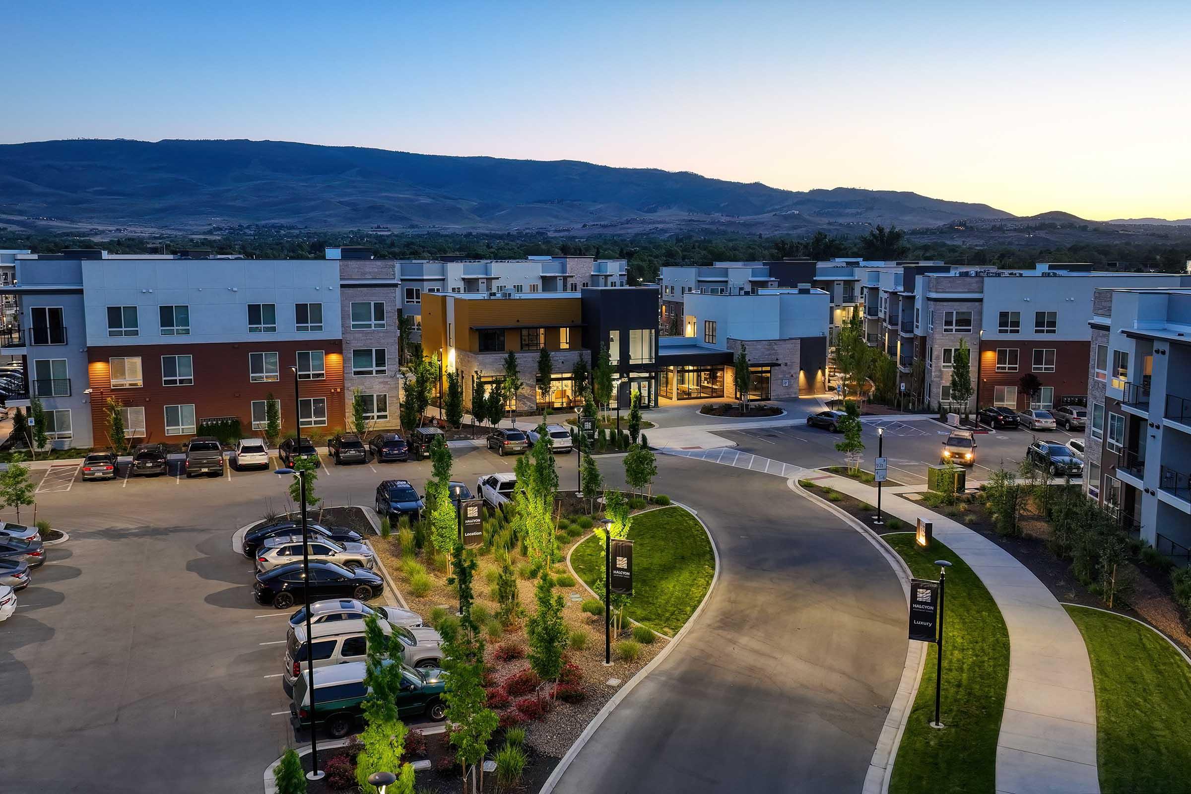 Aerial view of a modern apartment complex at dusk, featuring multiple three-story buildings, landscaped parking areas, and a welcoming entrance. The surrounding mountains and twilight sky create a serene atmosphere. The scene showcases well-maintained greenery and vehicles parked along the circular drive.