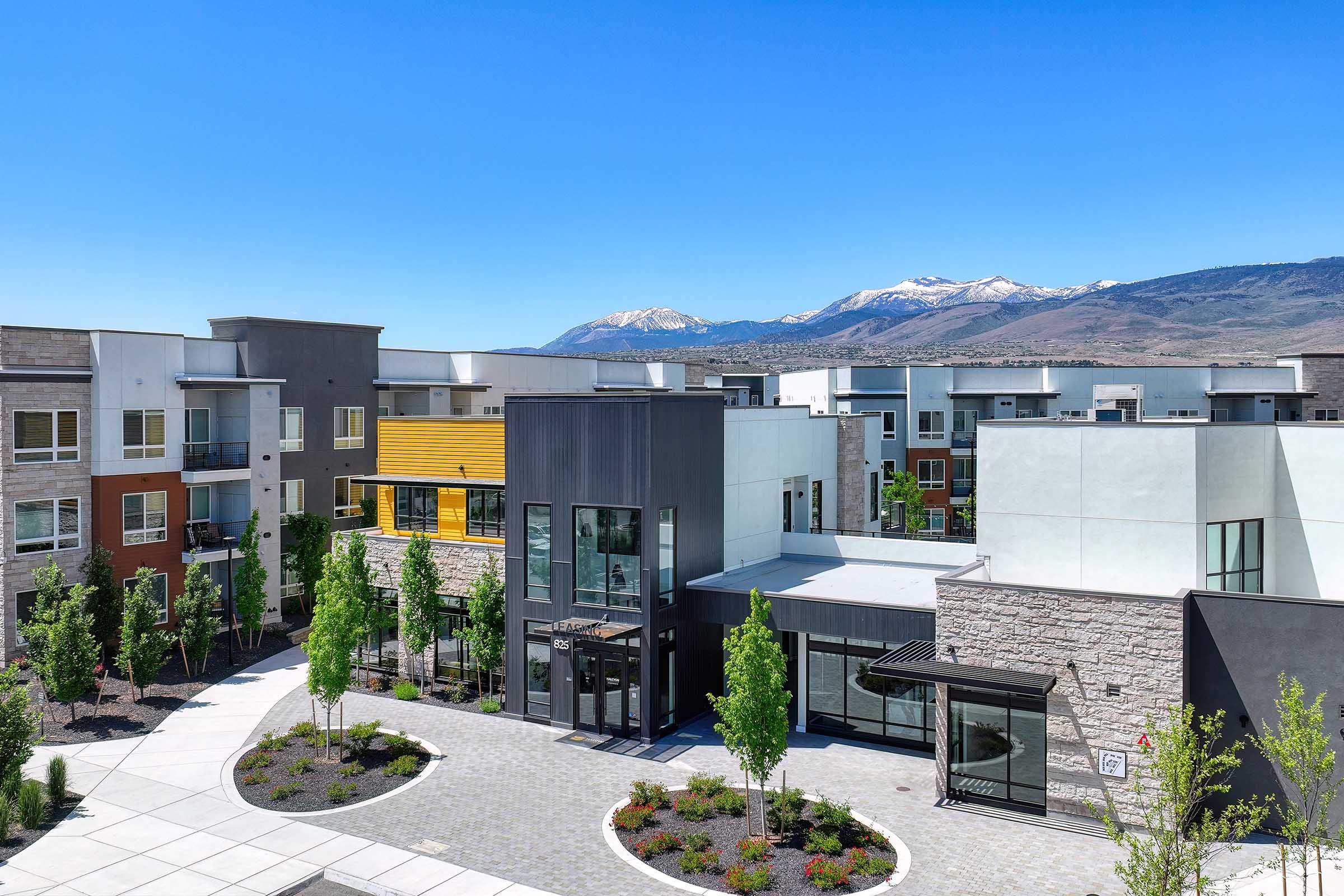A modern residential complex featuring contemporary buildings with varying facades, surrounded by landscaped greenery. The background showcases snow-capped mountains under a clear blue sky, creating a picturesque setting.