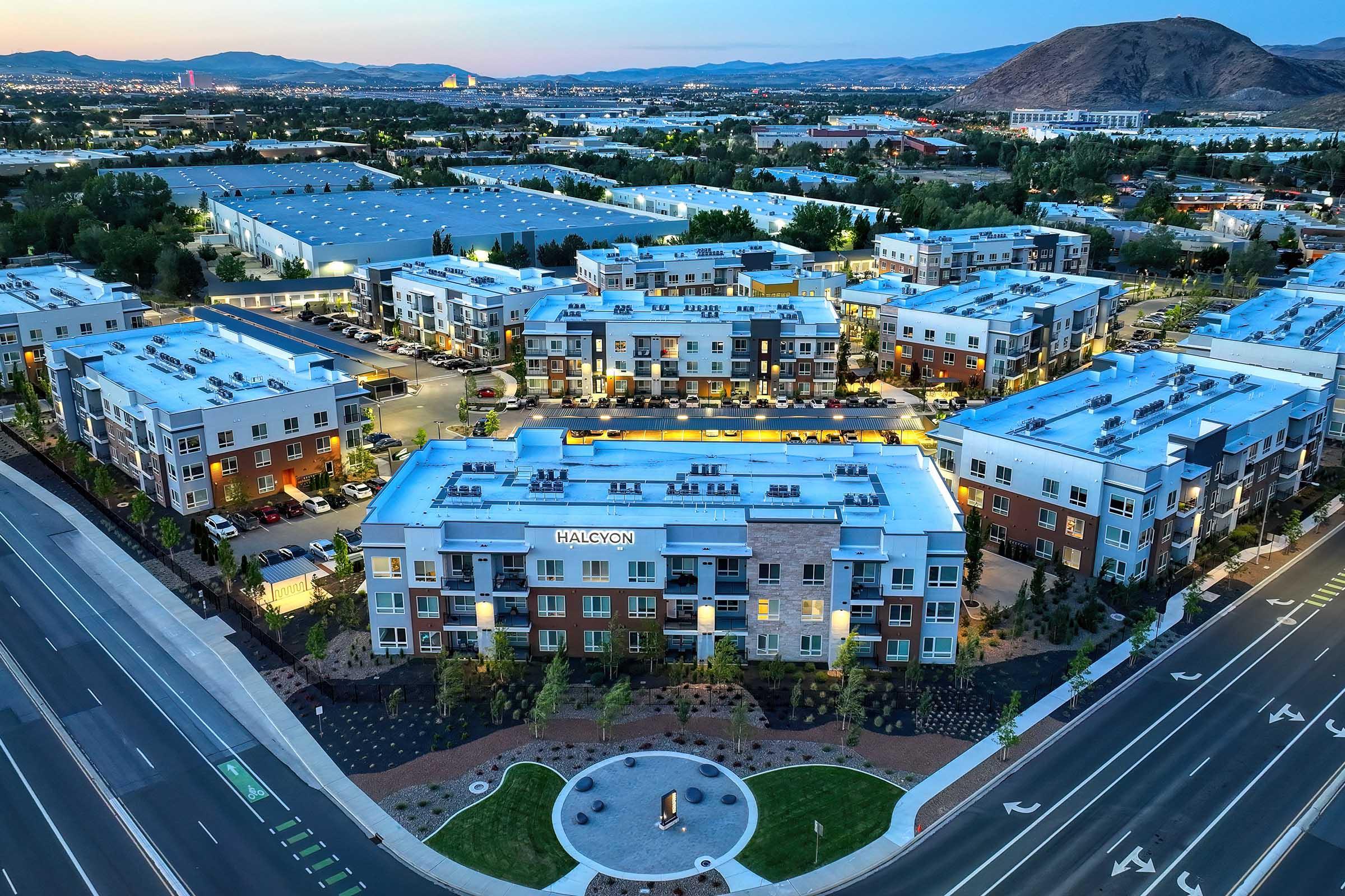 Aerial view of a modern residential complex named Halcyon, featuring multi-story buildings with flat roofs, surrounded by well-maintained landscaping. The complex is located near commercial buildings and open fields, with distant mountains in the background and clear skies, indicating a vibrant suburban area.