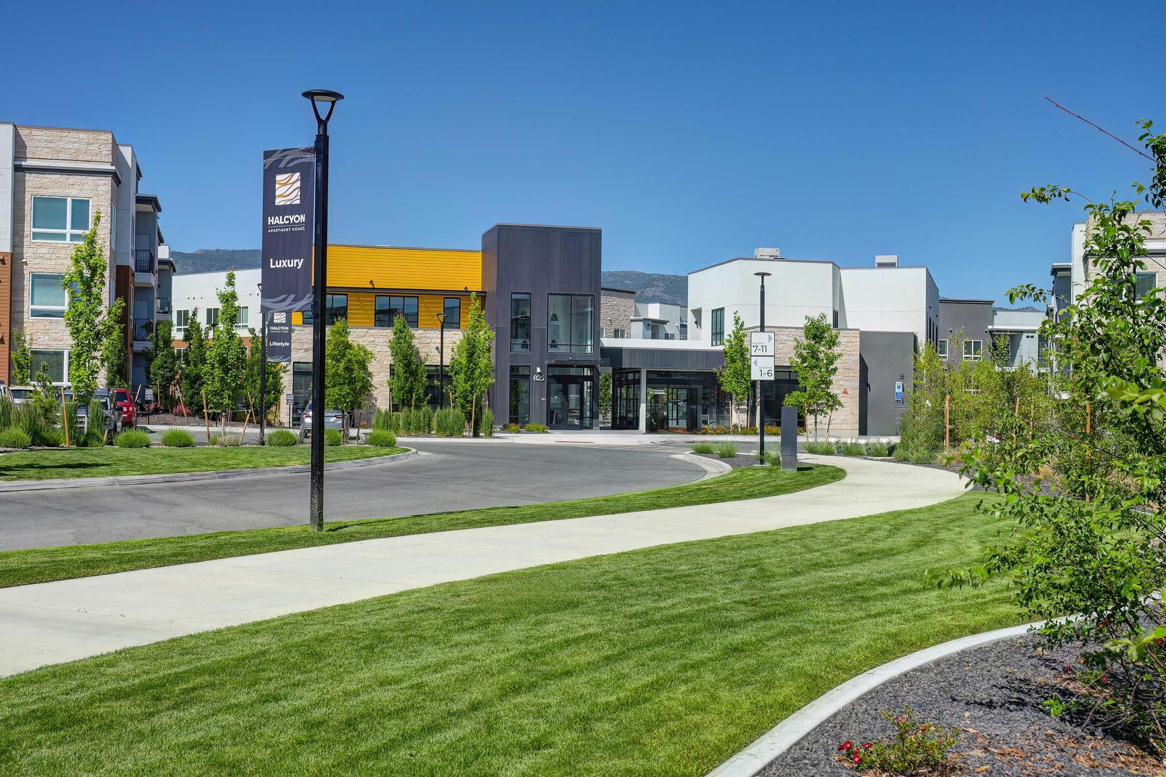 A modern residential community featuring sleek, contemporary architecture. The foreground includes a landscaped path and greenery, while the background showcases multiple buildings with a mix of colors, including gray and yellow facades. Clear blue skies create a bright, inviting atmosphere.