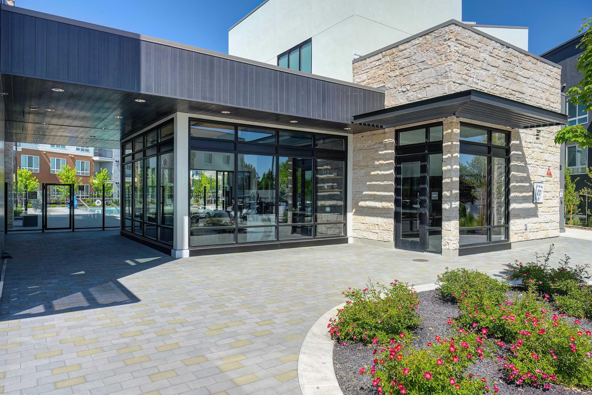Modern building exterior featuring a combination of stone and glass architecture. The entrance has large glass windows, allowing natural light to flood the interior. A landscaped area with blooming flowers and paved pathways enhances the inviting atmosphere, set against a clear blue sky.