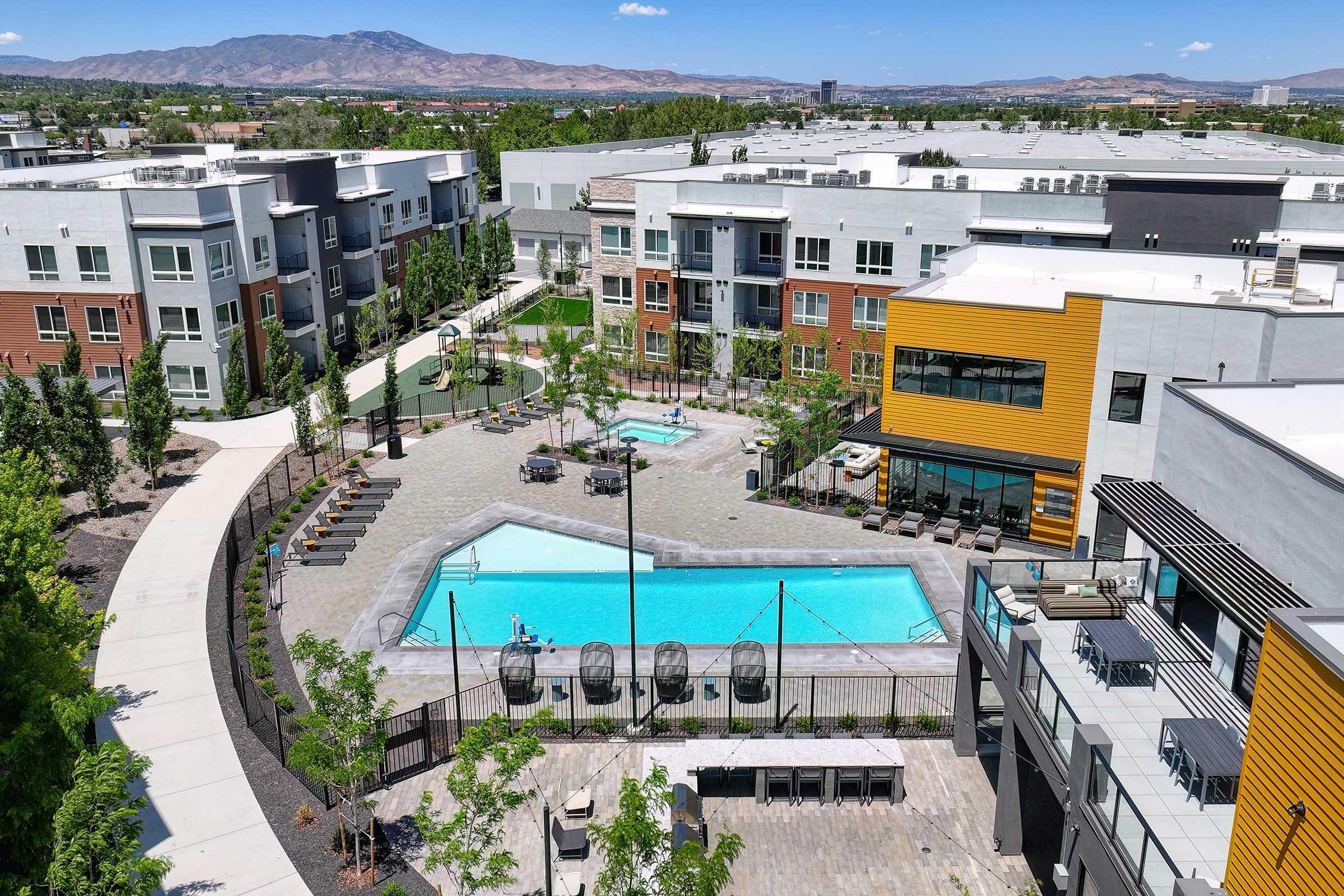 Aerial view of a modern apartment complex featuring a swimming pool surrounded by lounge chairs, spacious outdoor areas, and landscaped greenery. The buildings are a mix of colors, with a backdrop of mountains and blue skies, creating a vibrant, inviting atmosphere.