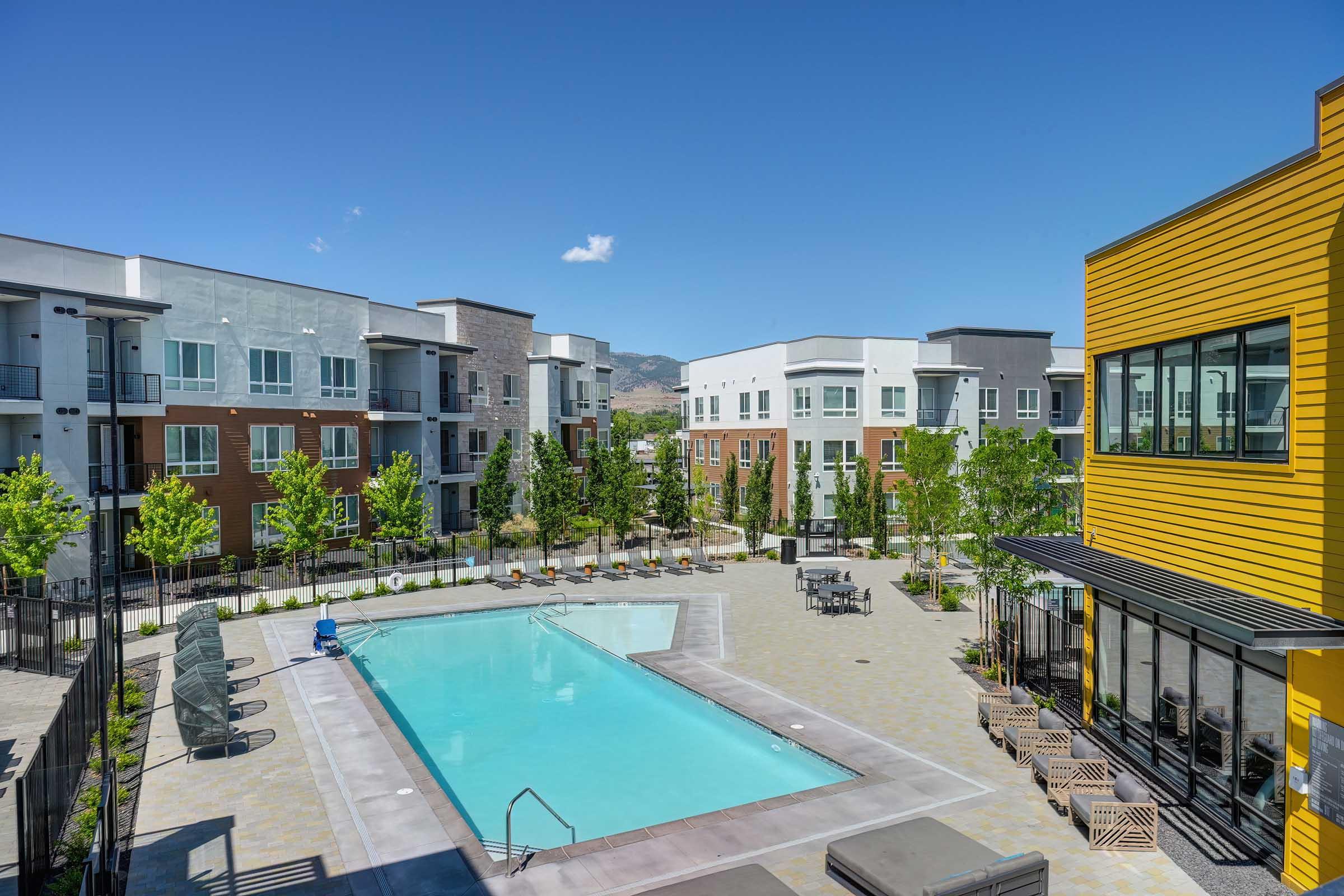 Modern apartment complex featuring a pool surrounded by lounge chairs and landscaped greenery. Bright blue sky above, with multiple buildings showcasing contemporary architecture in the background.