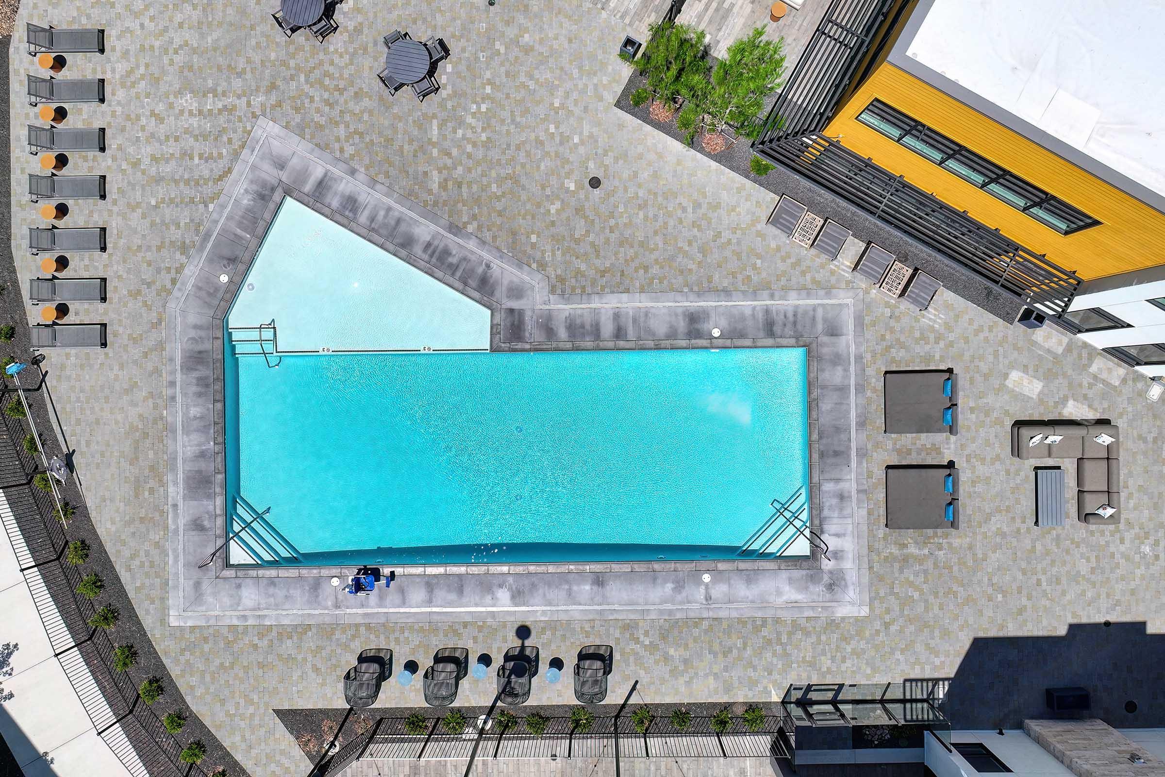Aerial view of a modern swimming pool surrounded by lounge chairs and tables. The pool features a unique shape with clear blue water, nestled within a landscaped area of an apartment complex. Sunlight reflects on the water's surface, creating a inviting atmosphere.