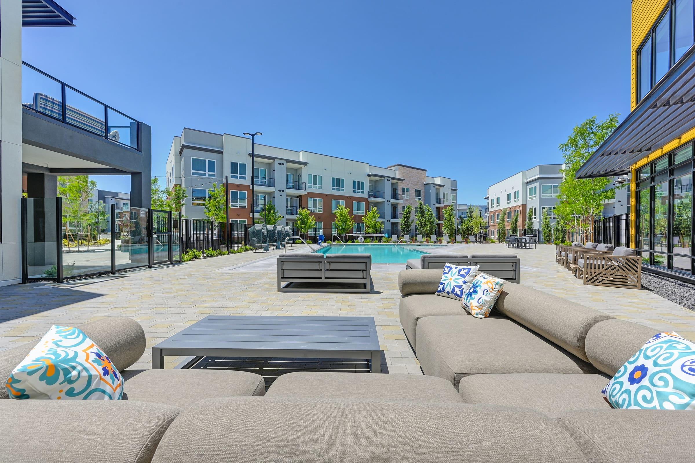 A modern outdoor lounge area featuring a large sectional sofa with colorful decorative pillows, facing a swimming pool surrounded by contemporary apartment buildings. Sunlight illuminates the space, highlighting landscaped greenery and patio furniture in a relaxed setting.