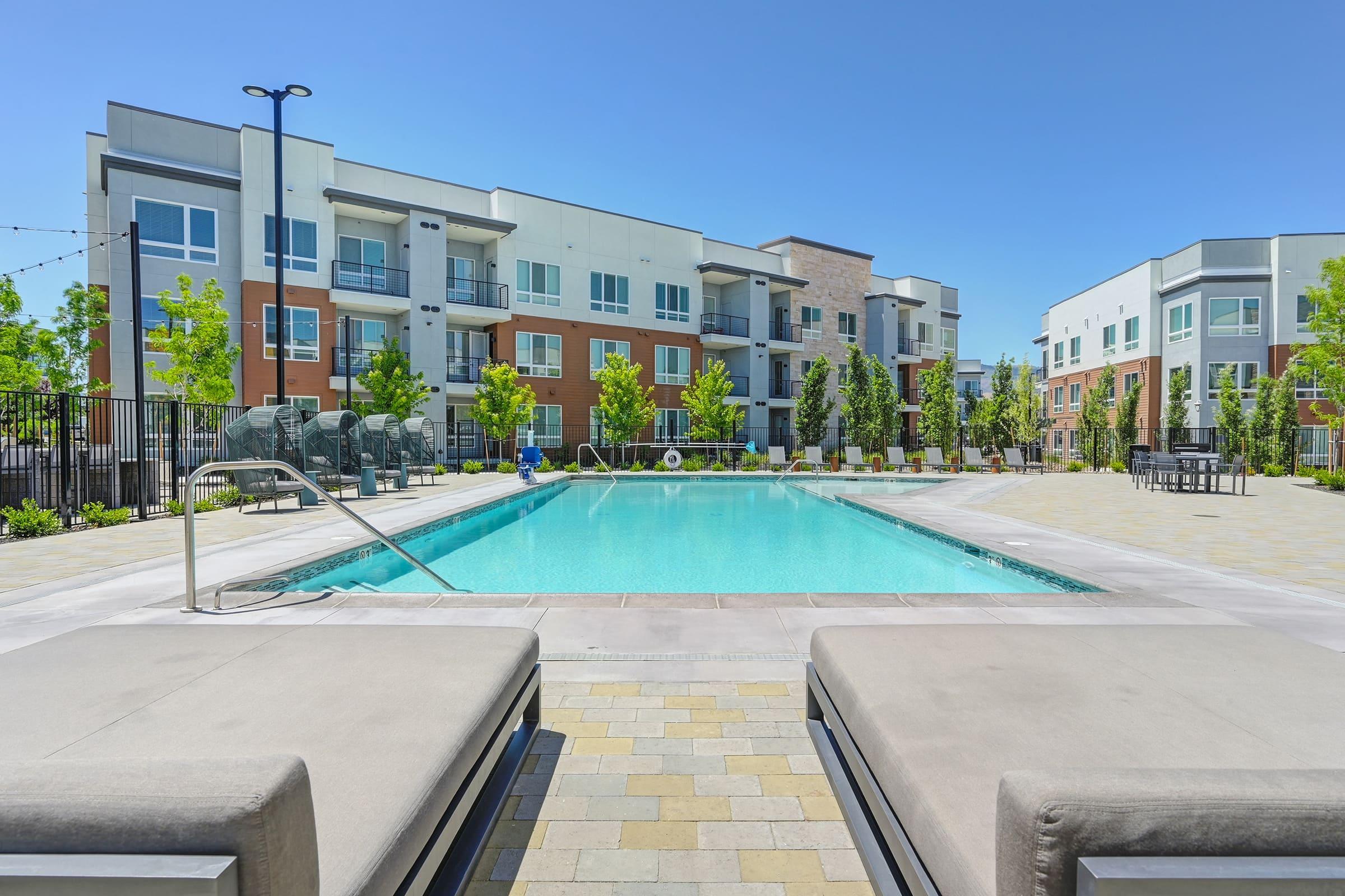 A modern apartment complex with a sparkling pool in the foreground. Lounge chairs are placed by the poolside, surrounded by well-maintained landscaping featuring trees and shrubs. The buildings in the background are contemporary, with balconies and large windows, set under a clear blue sky.