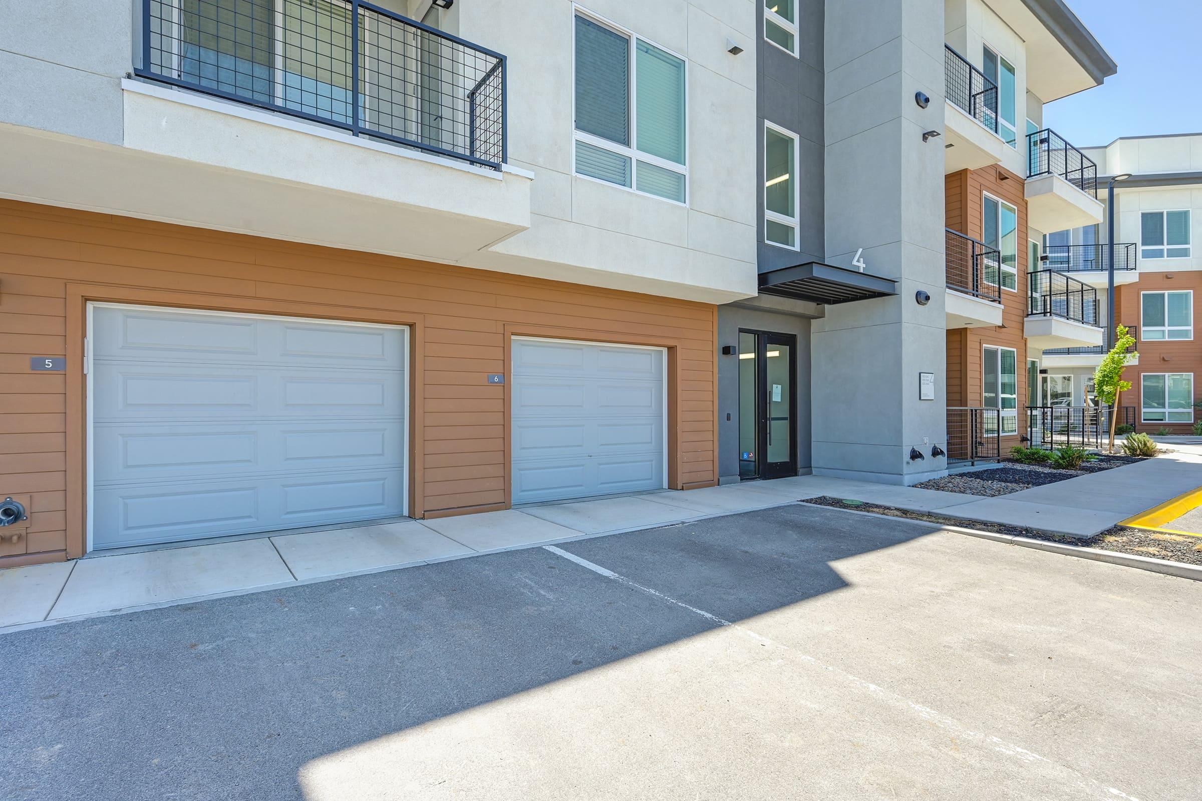 A modern apartment building featuring two garage doors, one of which is labeled '4', with a well-maintained entrance area and nearby parking spaces. The exterior showcases a mix of light-colored siding and brick materials, with large windows providing natural light.