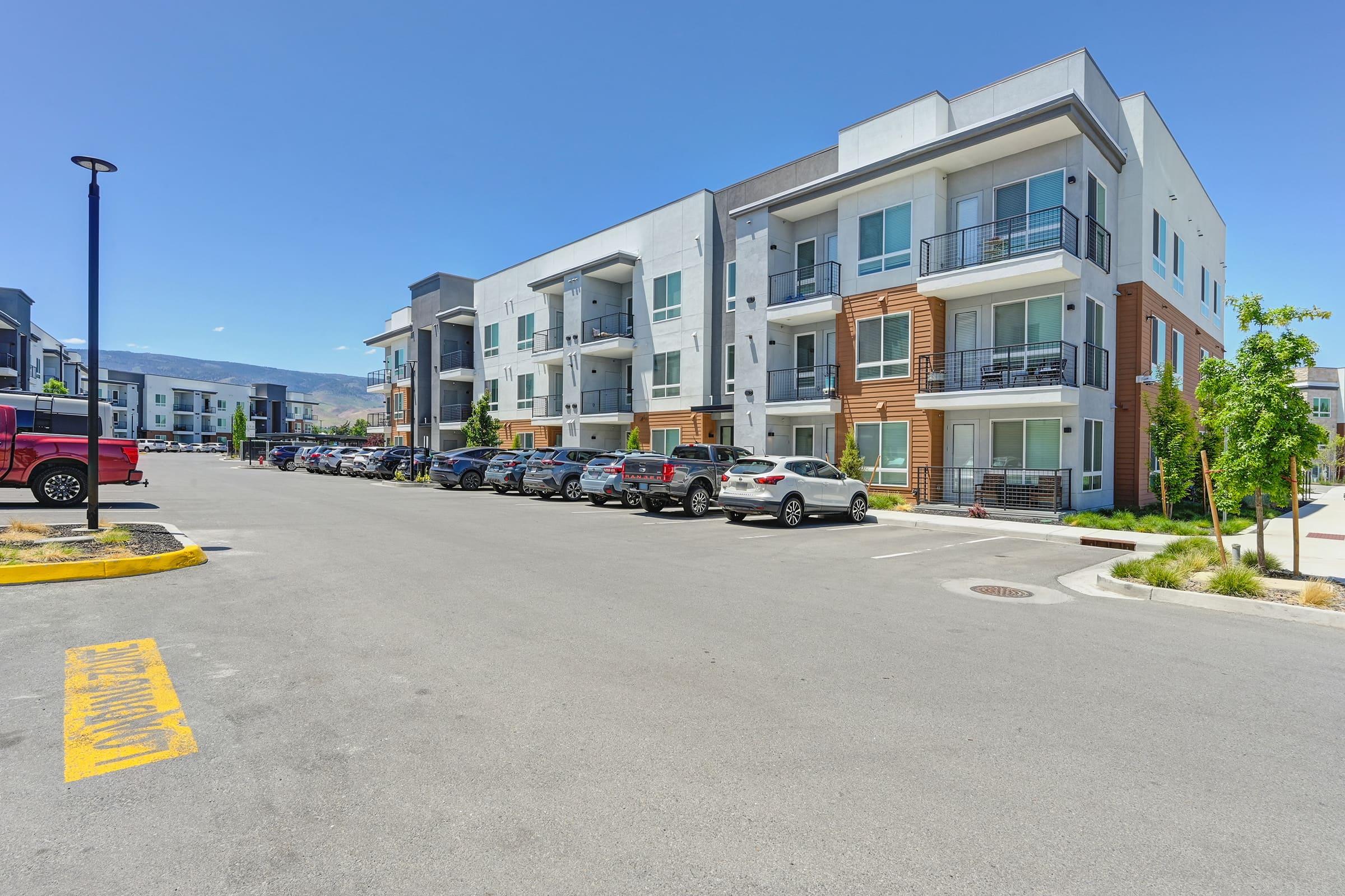 A modern residential apartment complex featuring three-story buildings with a mix of light and dark exteriors. The foreground shows a parking lot filled with various cars, and clean landscaping with trees and shrubs. Clear blue sky and bright sunlight enhance the vibrant look of the area.