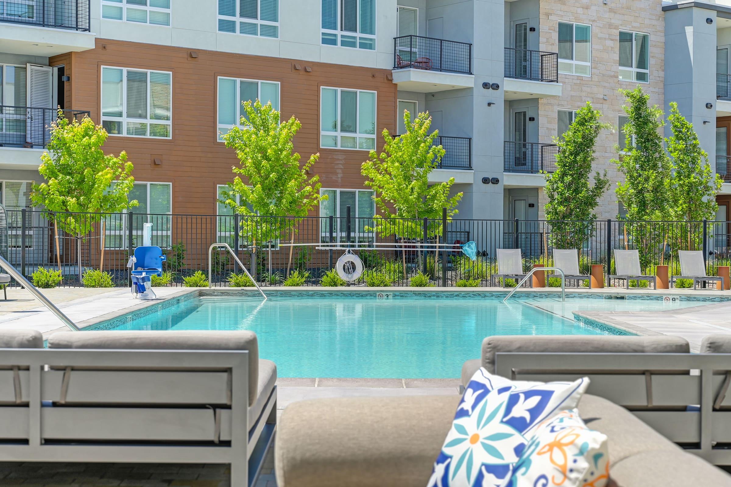 A sunny outdoor pool area surrounded by modern apartment buildings. Lush green trees line the space, and several lounge chairs are arranged nearby. In the foreground, there is a cozy seating area with a decorative pillow, adding to the inviting atmosphere of relaxation.