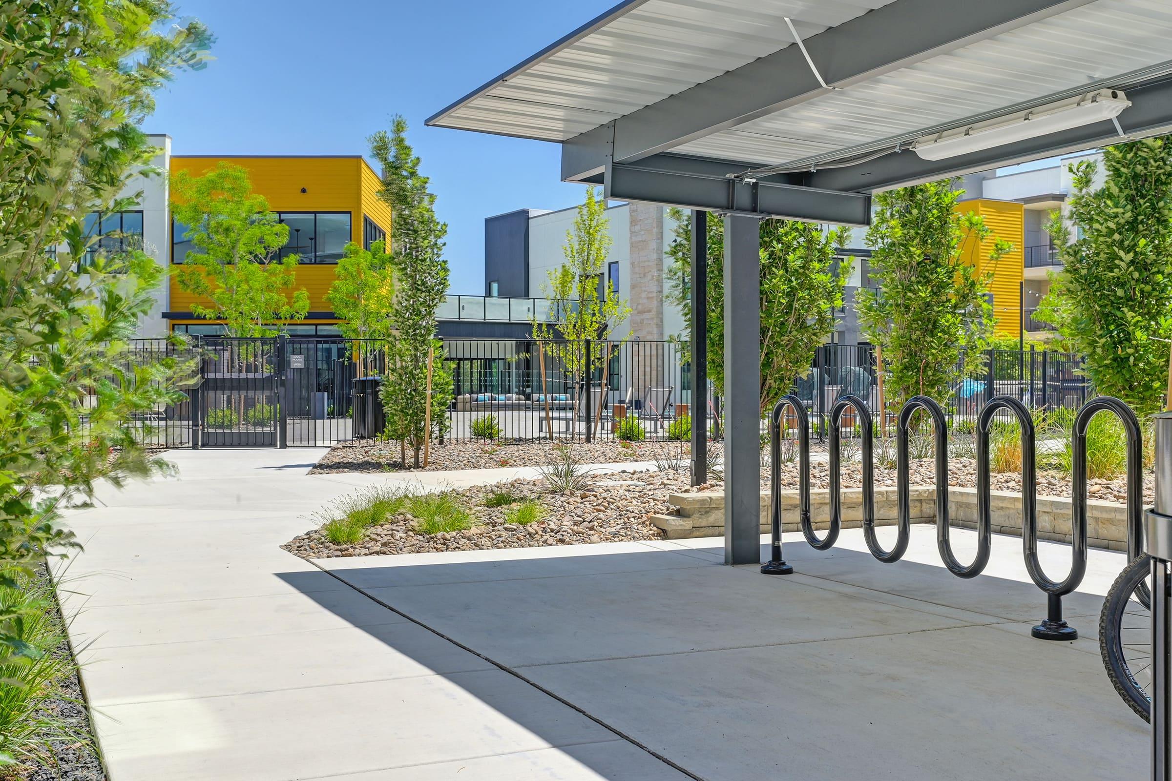 A modern outdoor bike rack beneath a canopy, with landscaped greenery and plants nearby. In the background, brightly colored yellow and gray buildings are visible, along with a fenced area. The scene is bright and sunny, highlighting a contemporary urban environment.