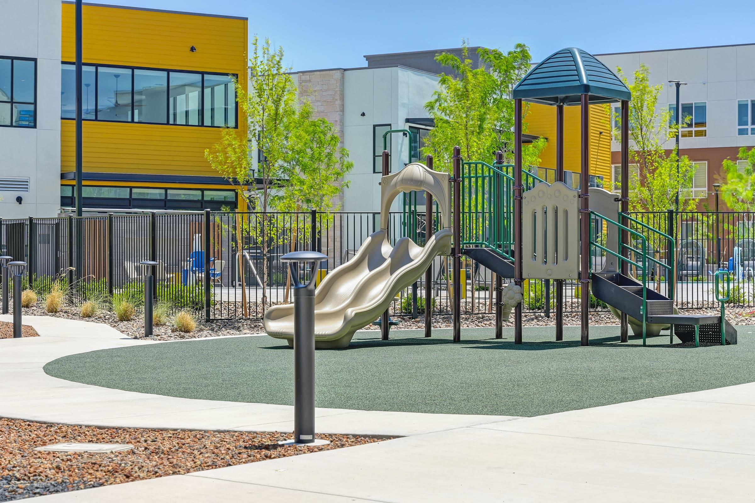 A modern playground featuring a green and yellow play structure with a slide, surrounded by grass and decorative rocks. The background includes contemporary buildings and trees, creating a bright and inviting outdoor space ideal for children to play.