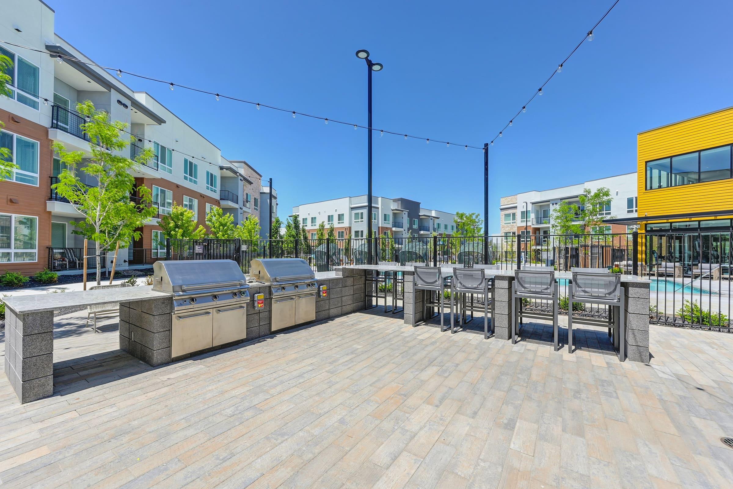 Outdoor communal grilling area with modern barbecue grills and bar-style seating, surrounded by landscaped greenery and contemporary apartment buildings. String lights overhead add ambiance. Clear blue sky enhances the inviting atmosphere.