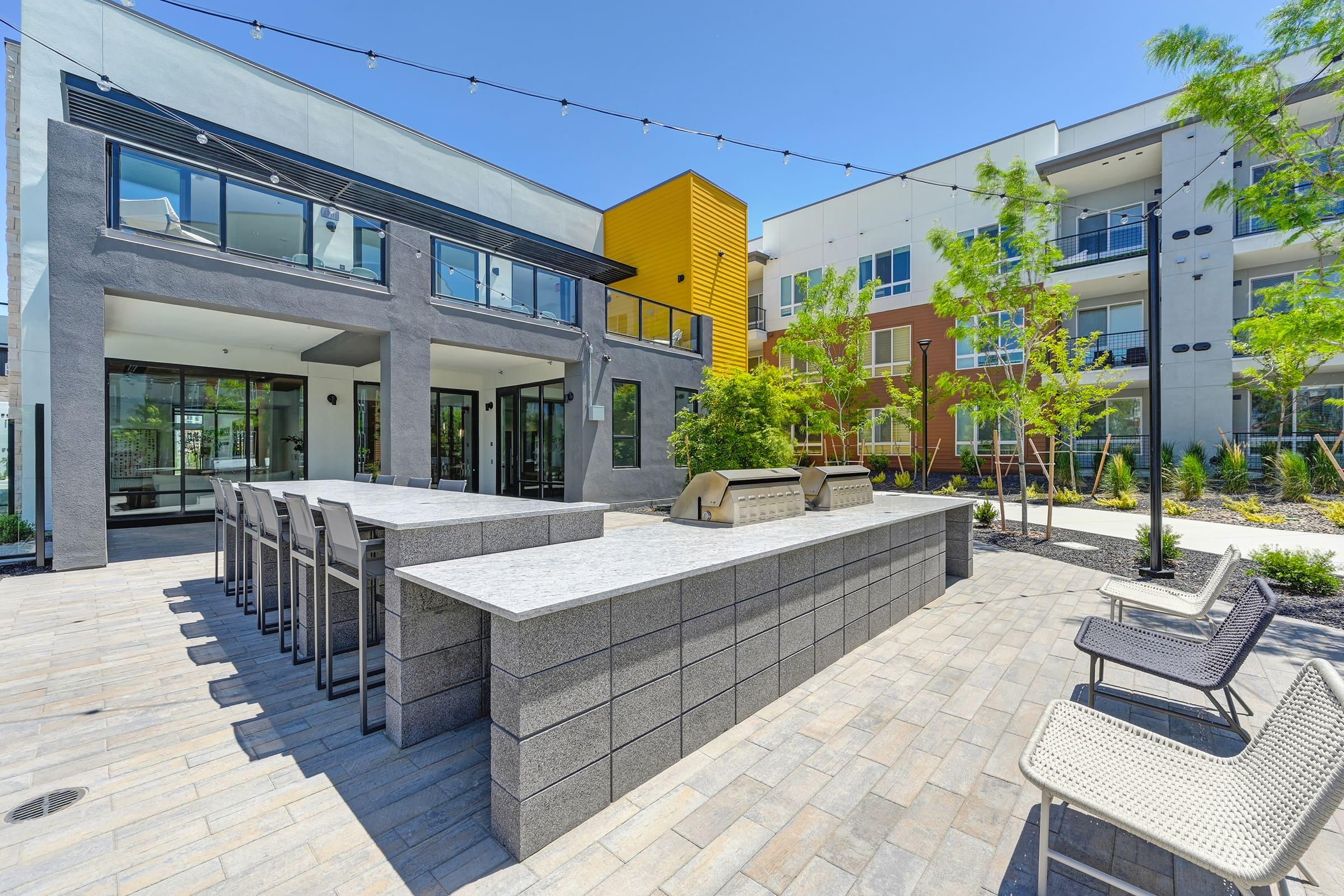 Modern outdoor lounge area featuring a sleek stone bar with high chairs, surrounded by trees and decorative landscaping. In the background, contemporary buildings with large windows and balconies are visible, creating a welcoming communal environment. String lights add a cozy touch to the space.