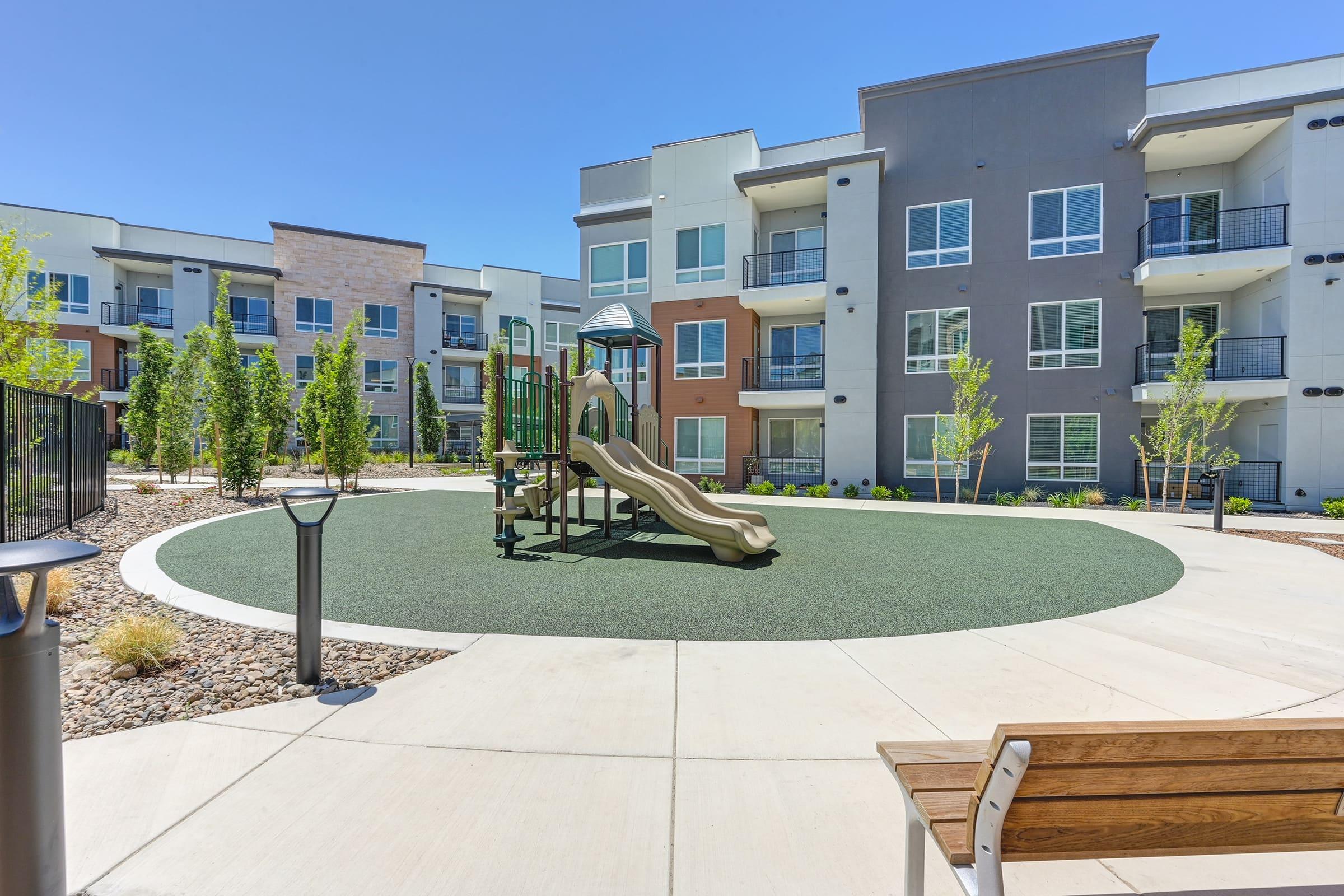 Playground with a slide and climbing structure in a landscaped area surrounded by modern apartment buildings. The setting features green grass-like surfacing, trees, and benches, under a clear blue sky, creating a welcoming environment for children and families.