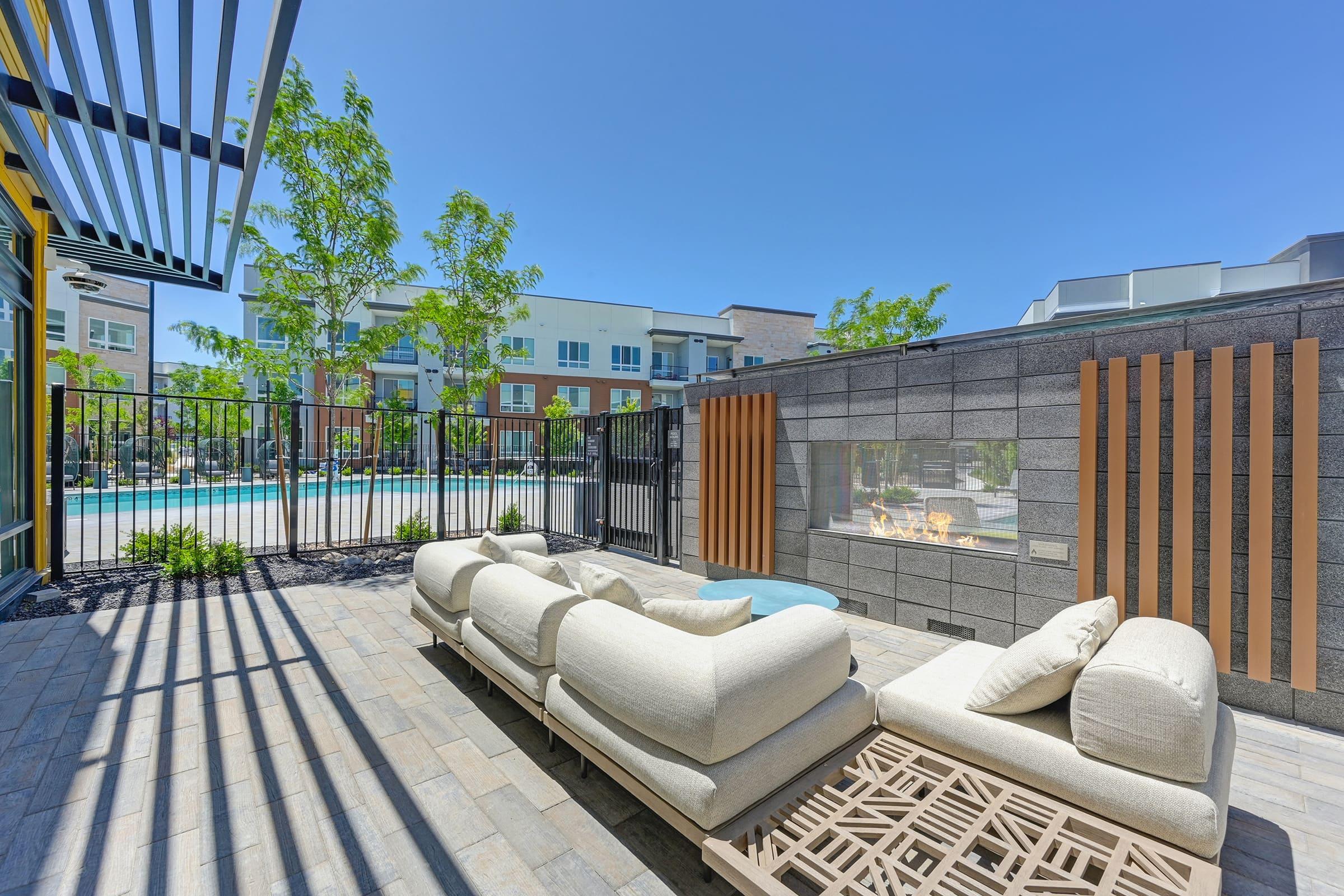 Outdoor lounge area featuring a stylish sectional sofa facing a modern stone wall with a fire feature. In the background, a swimming pool and landscaped greenery are visible under a clear blue sky, creating a relaxing atmosphere.