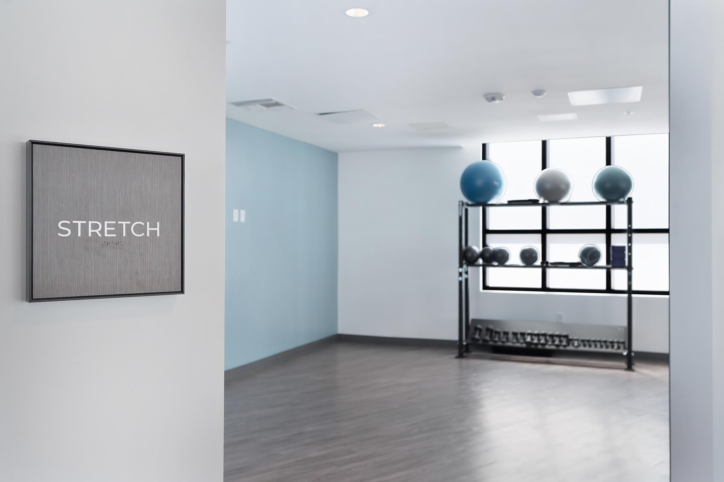 A modern fitness studio interior featuring a "STRETCH" sign on the wall. The room has a clean, minimalist design with light hardwood flooring. In the background, there is a shelf with exercise balls of various colors and a rack with dumbbells, creating a welcoming atmosphere for workouts.